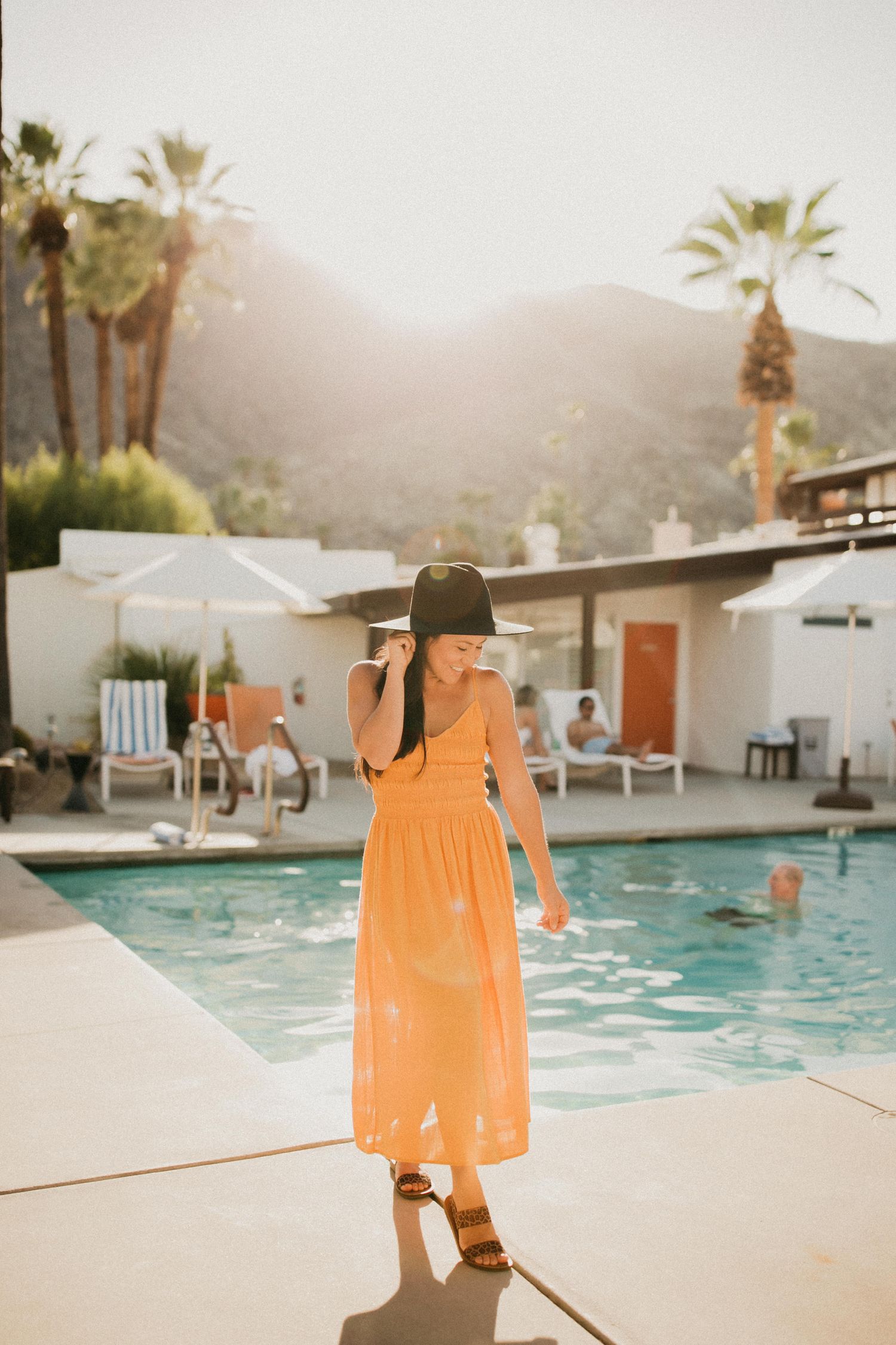 Woman in orange dress and sun hat standing by a palm-lined pool at sunset.