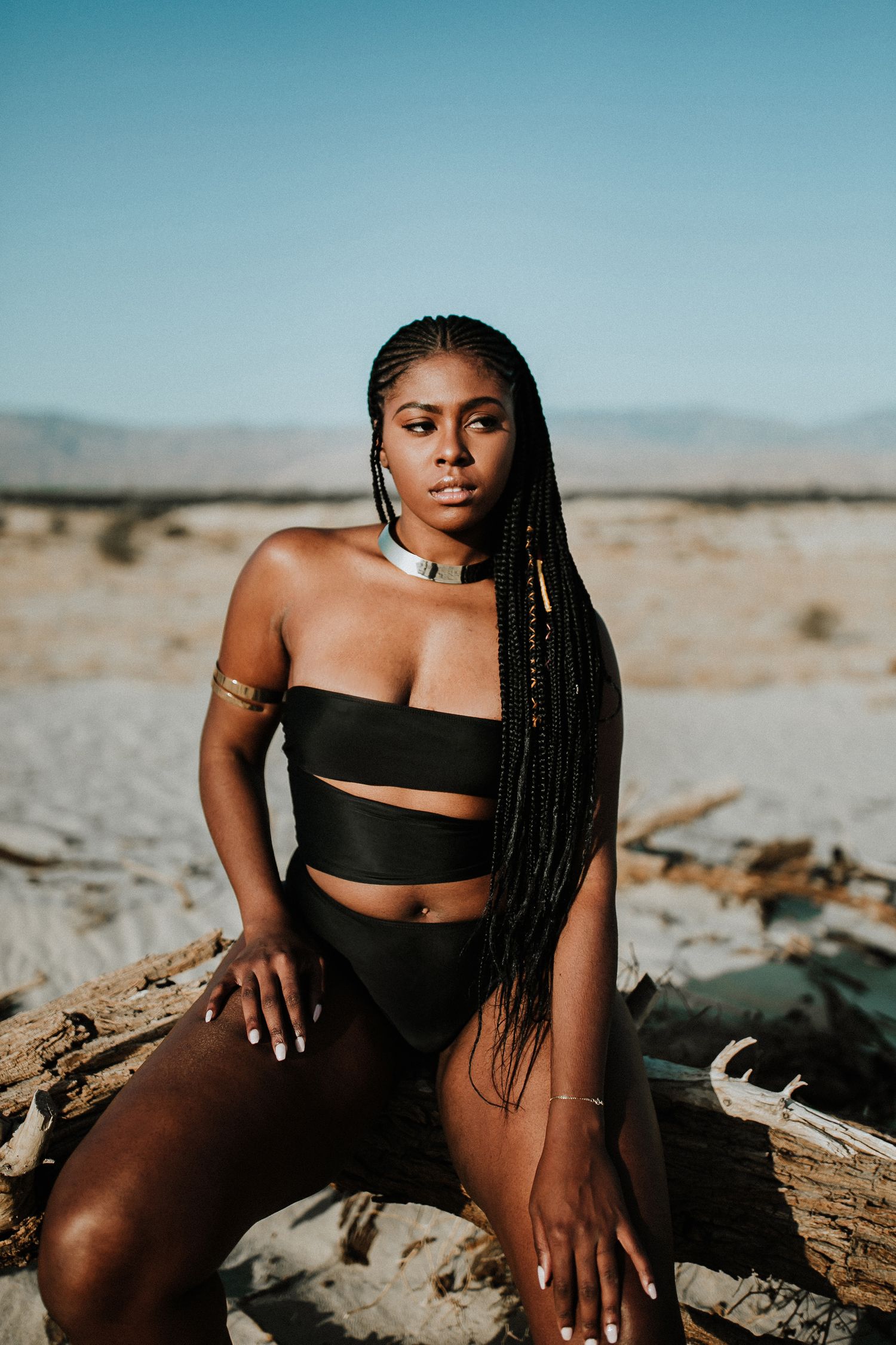 Woman with long braids poses in black swimwear on desert landscape.