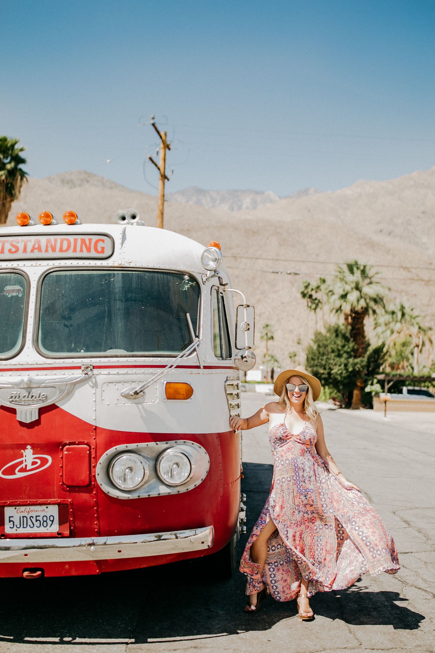 Vintage red and white bus parked in desert landscape with palm trees and mountains in background.