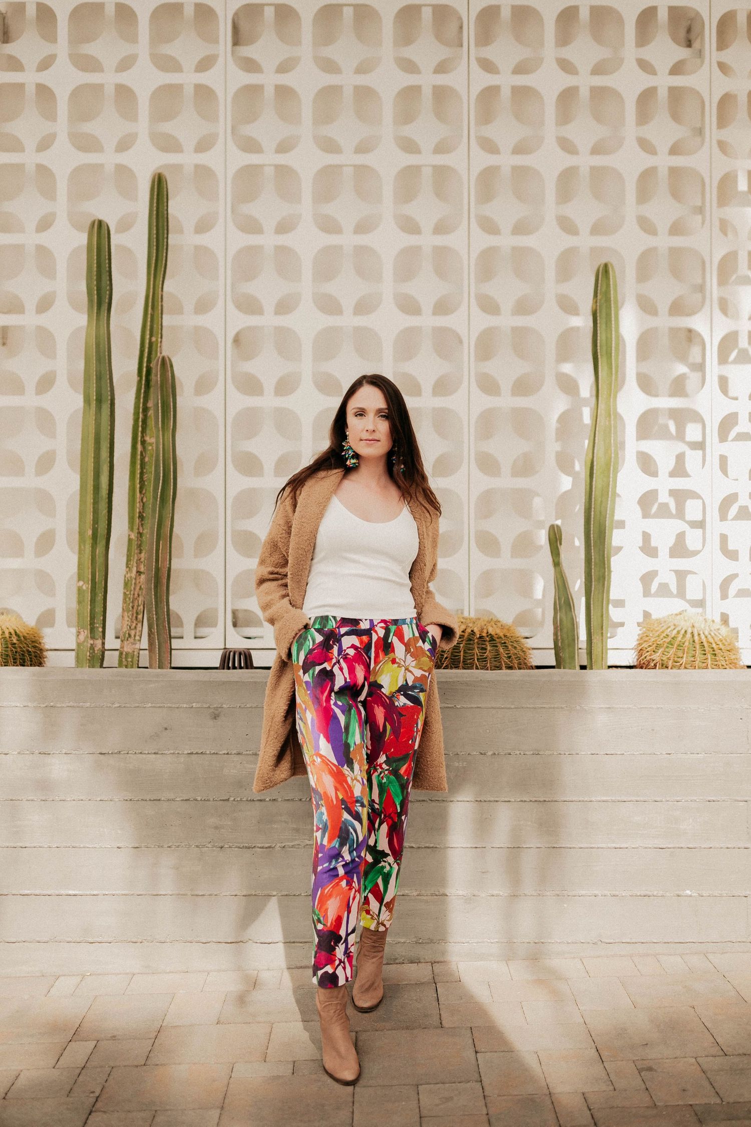 Stylish woman in colorful floral pants and tan cardigan poses against geometric wall with tall cacti.