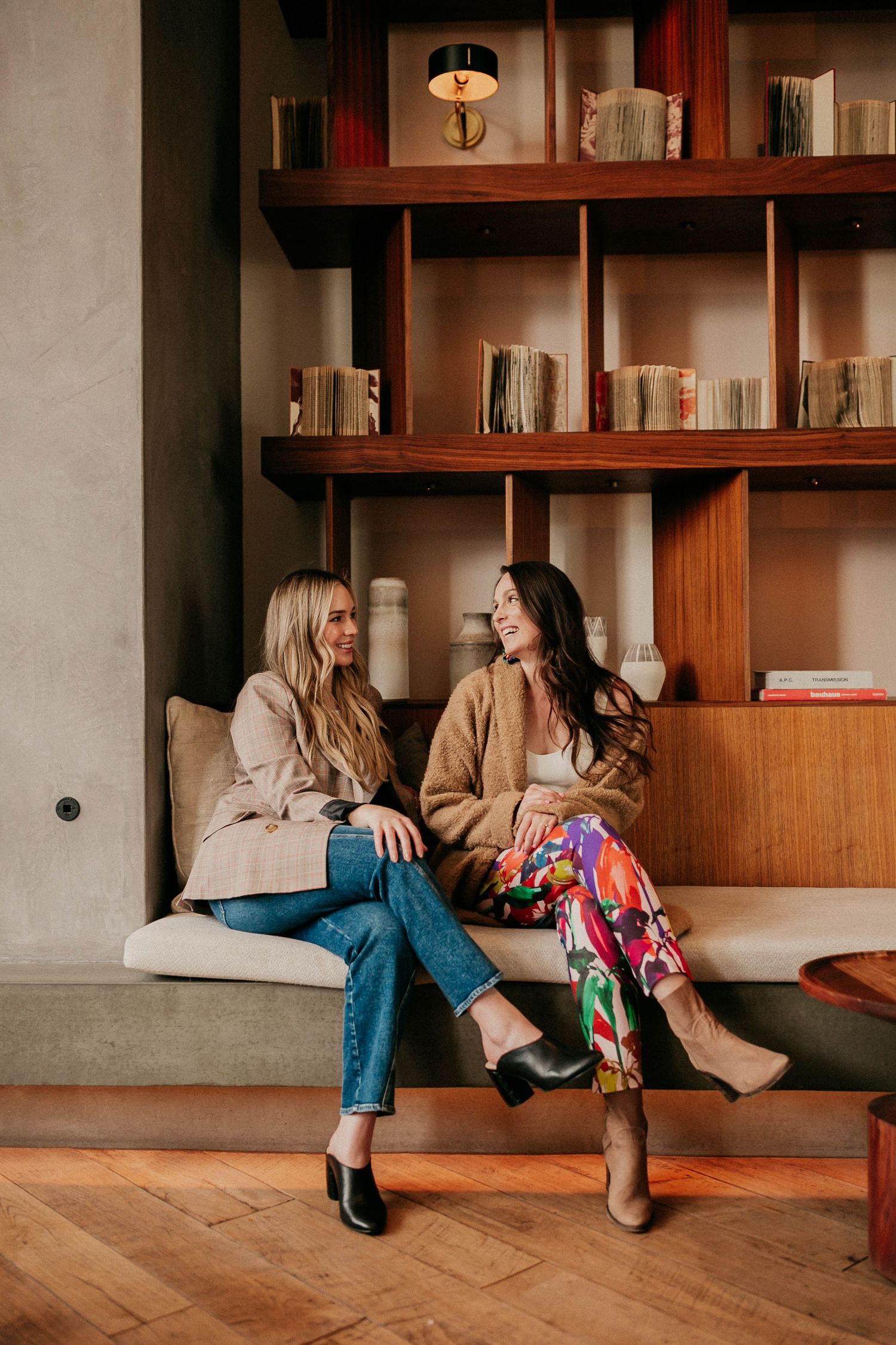 Two friends in casual outfits chat while sitting on couch against wooden bookshelf.