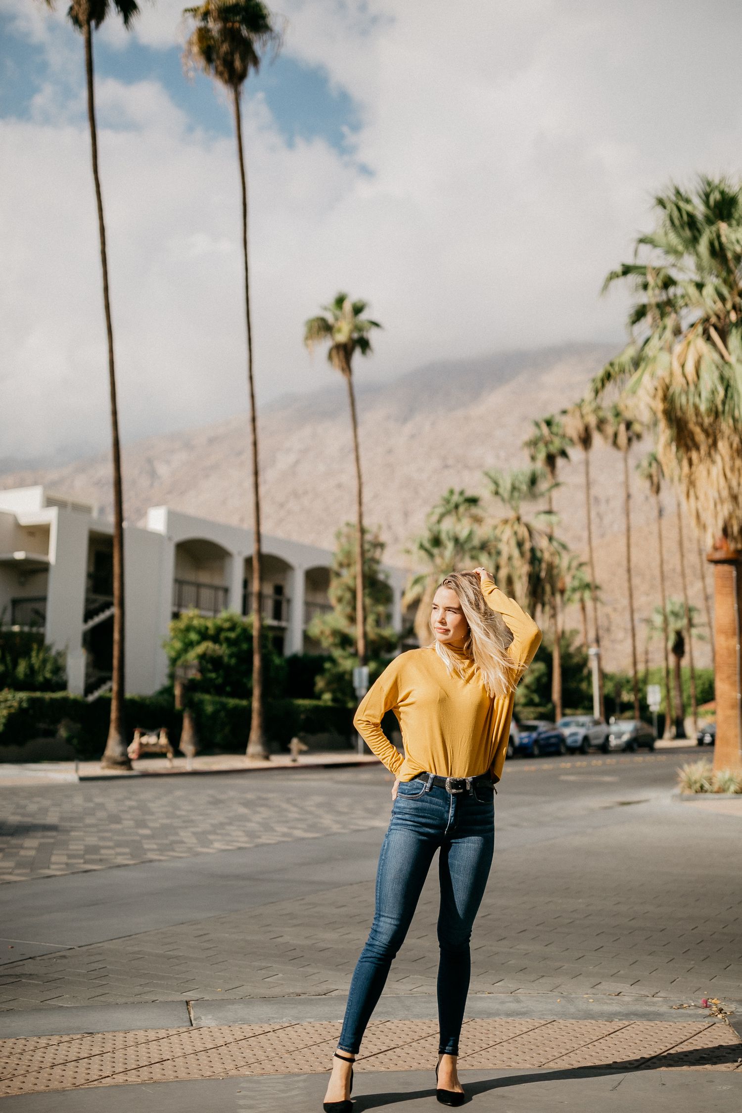 Woman in yellow top and jeans poses on a palm tree-lined street with mountains in the background.