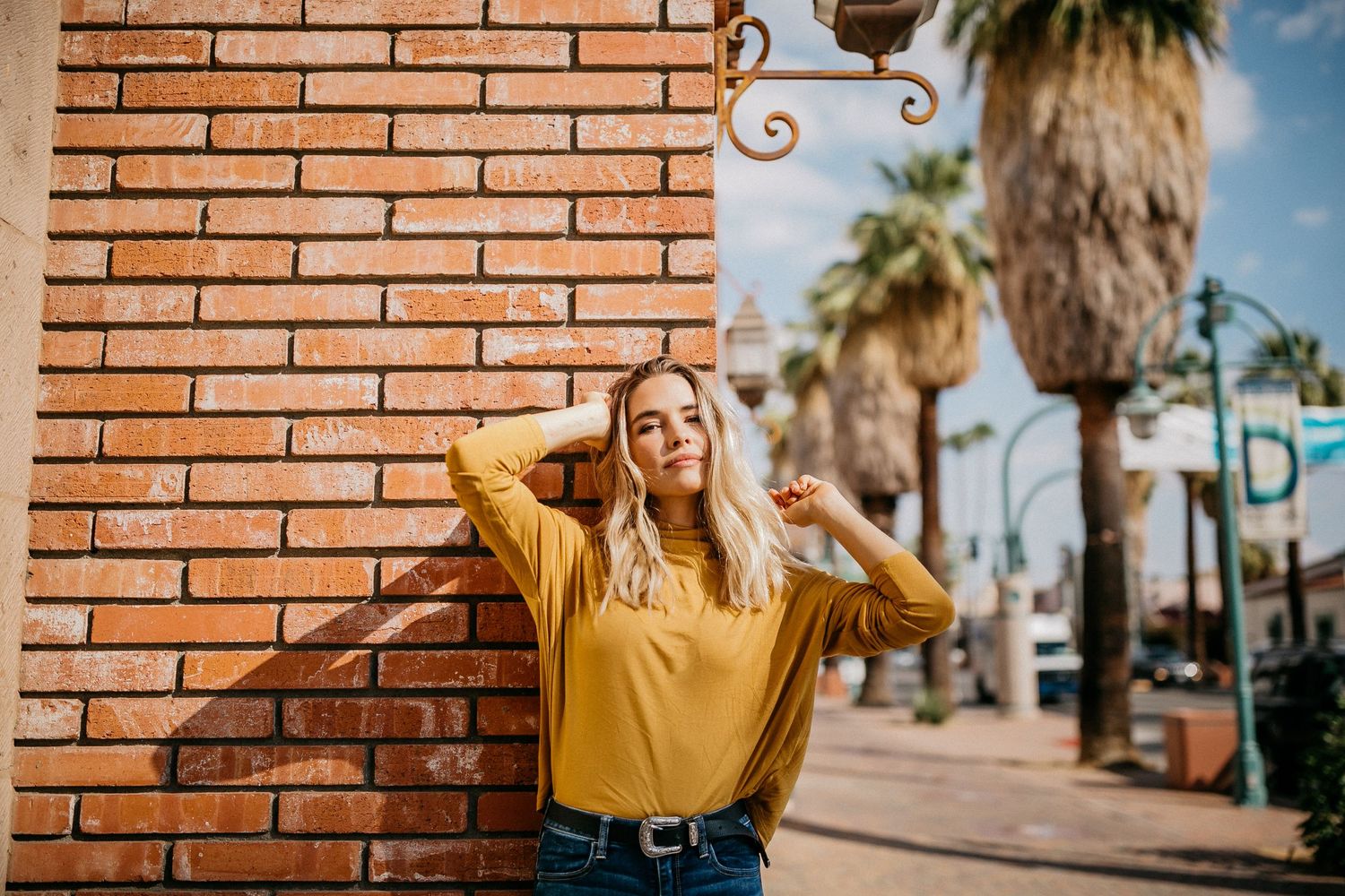 Woman in yellow top poses against brick wall with palm trees in background on sunny day.