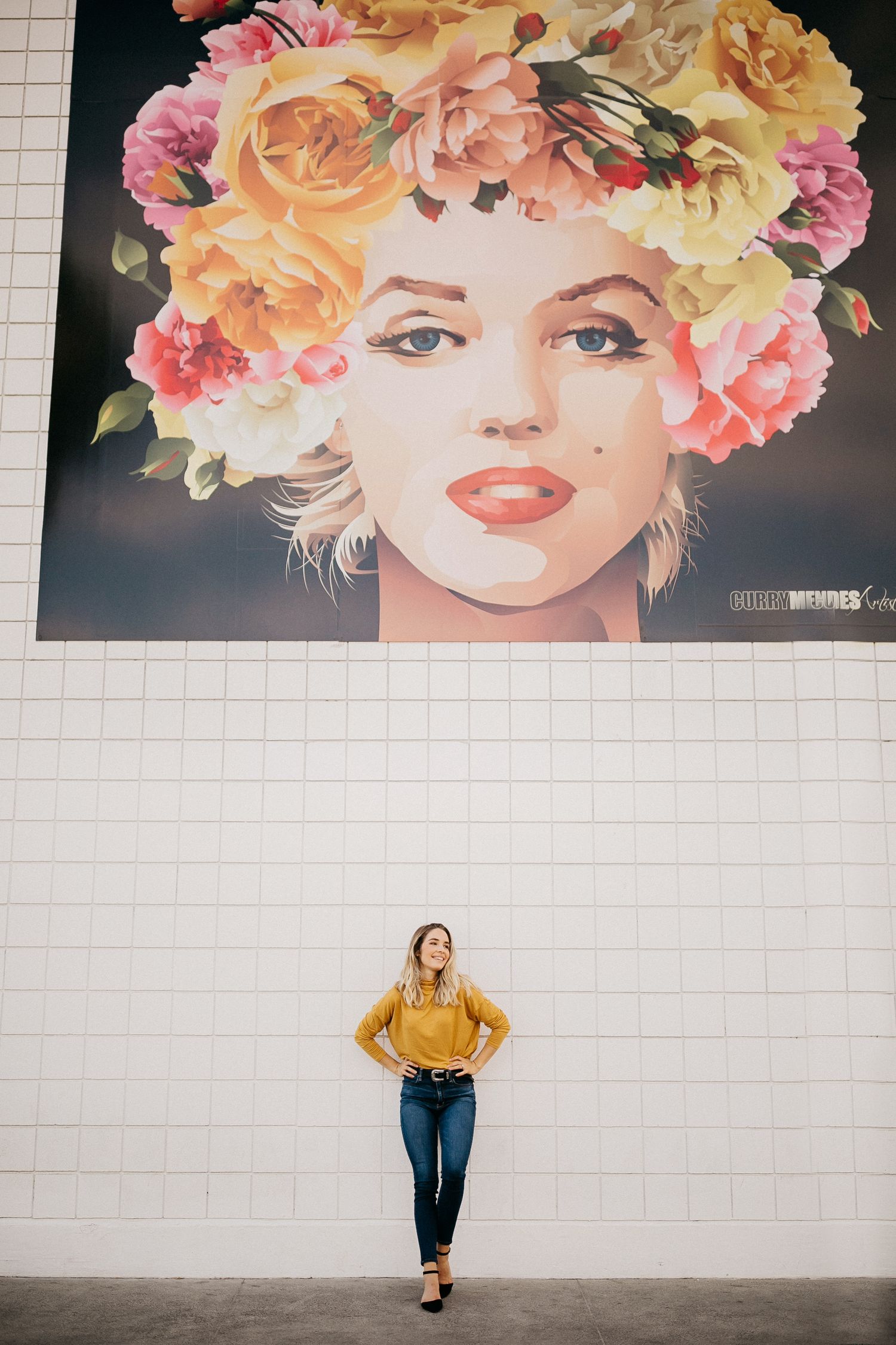 Woman in mustard yellow sweater poses against white wall beneath large floral portrait mural.