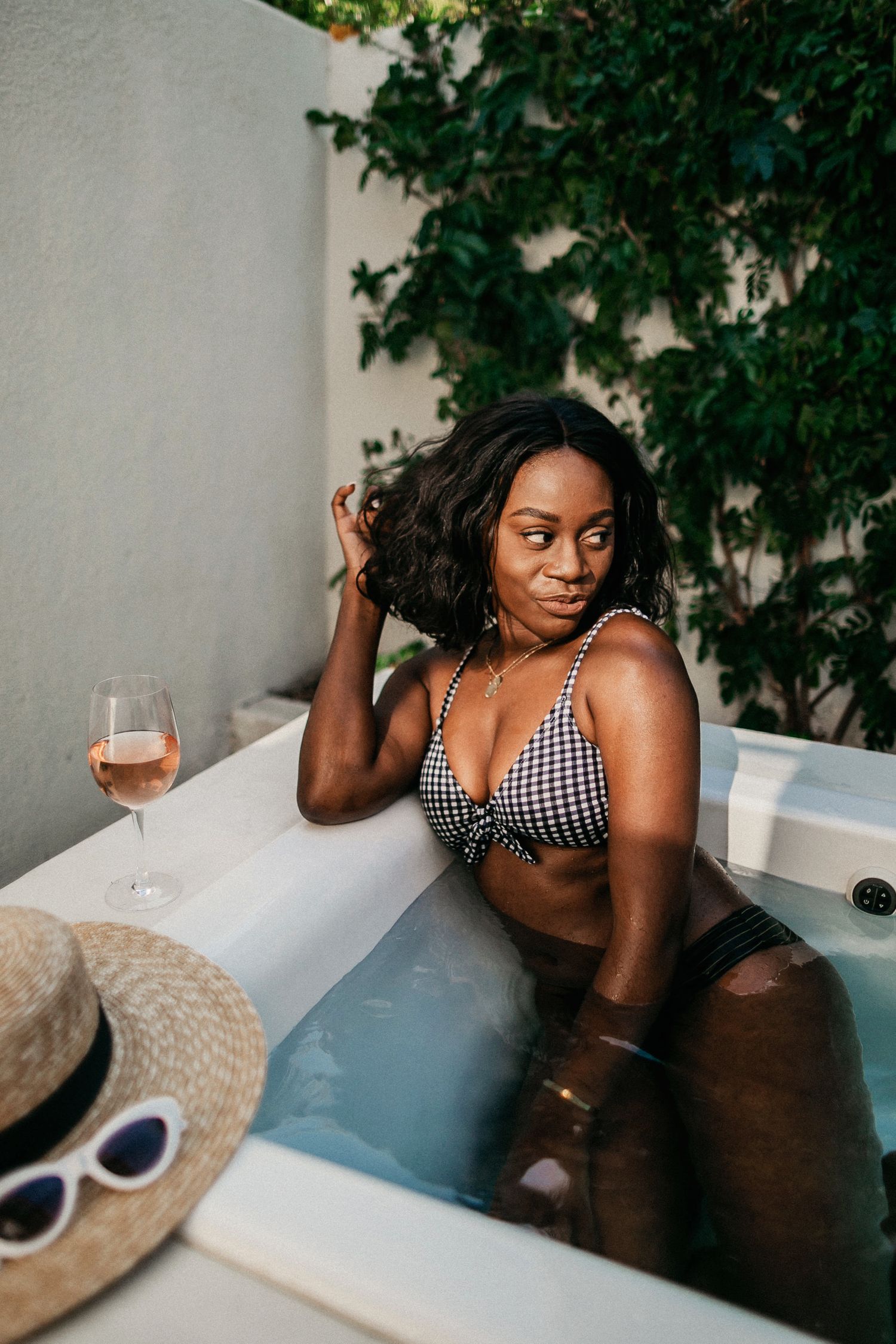 Woman relaxes in outdoor hot tub with wine glass and sun hat nearby.