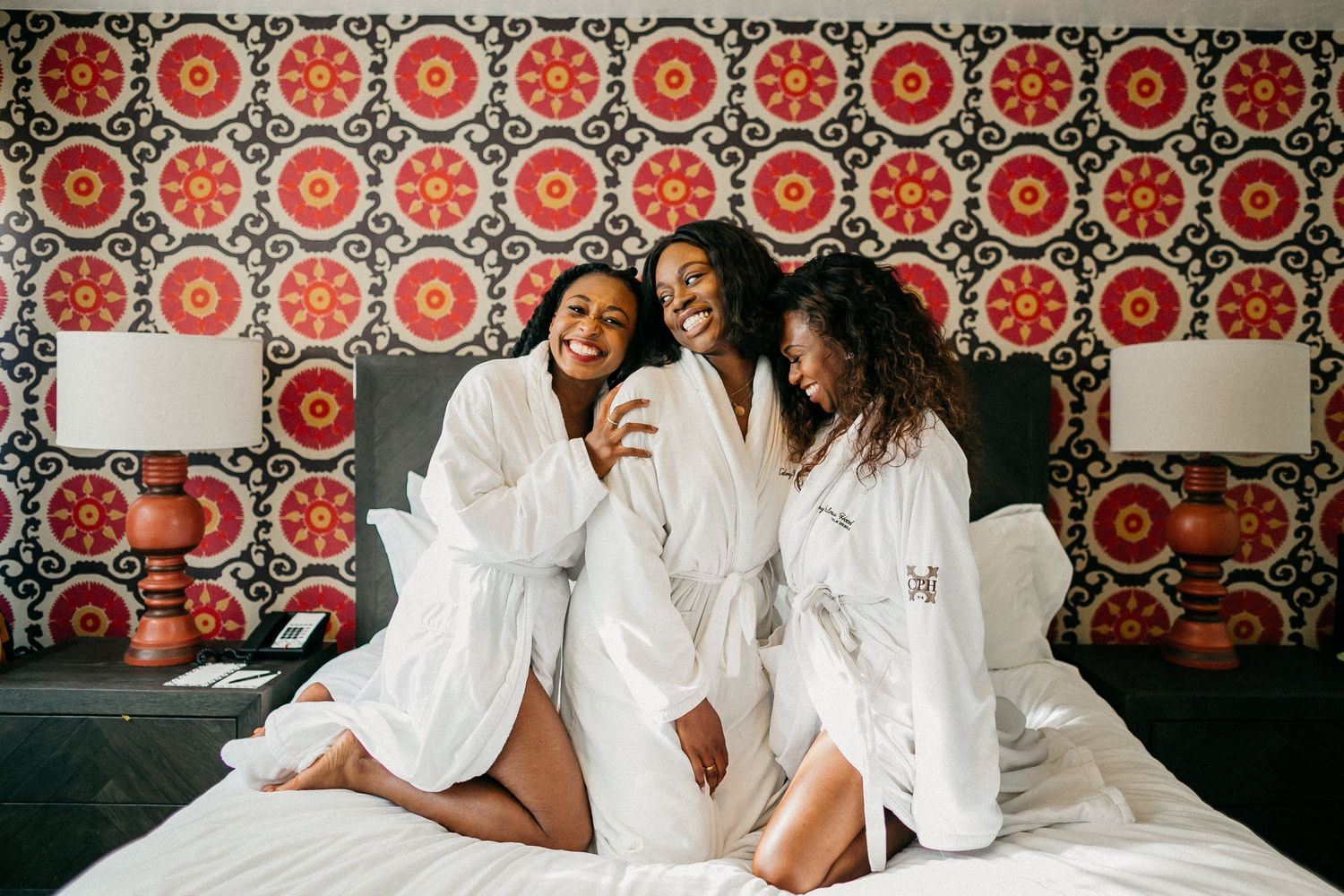 Three women in white robes laugh together on hotel bed with retro orange patterned wallpaper.