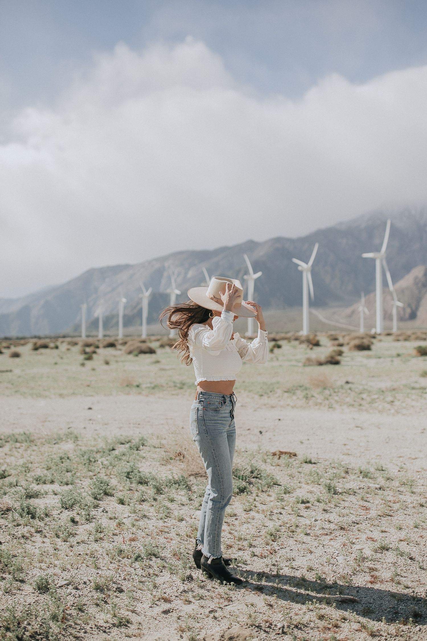 Wind turbines line distant mountains as figure in casual outfit stands in desert landscape.