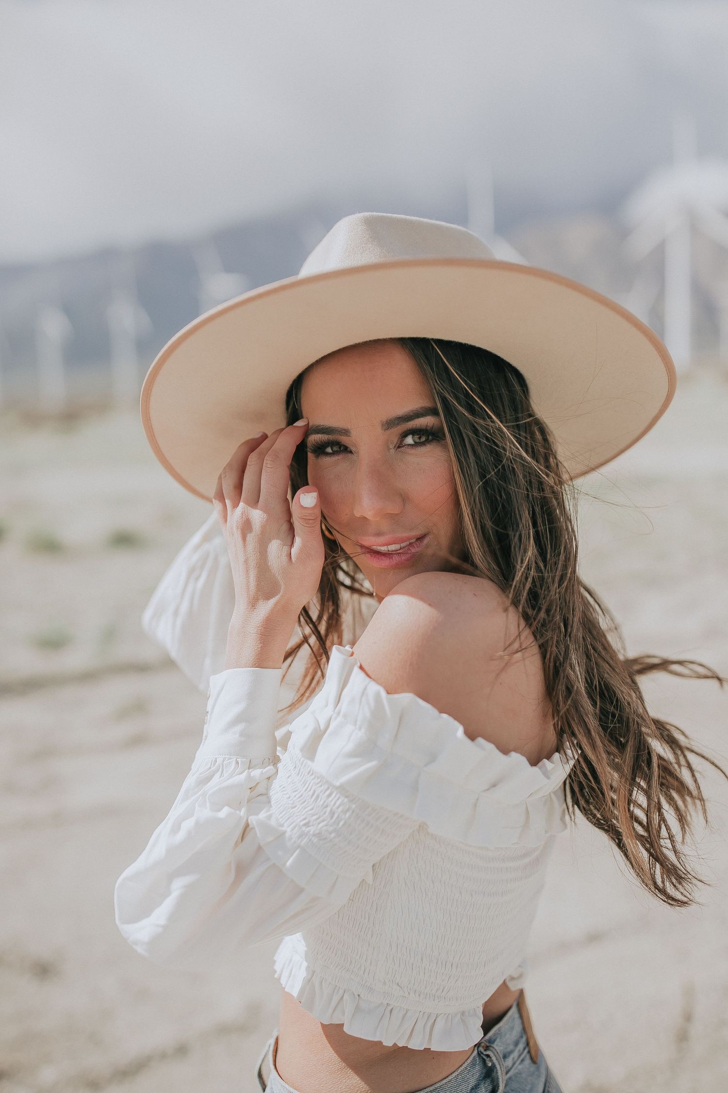 Desert fashion portrait with white off-shoulder top and wide brim hat against wind turbine landscape.
