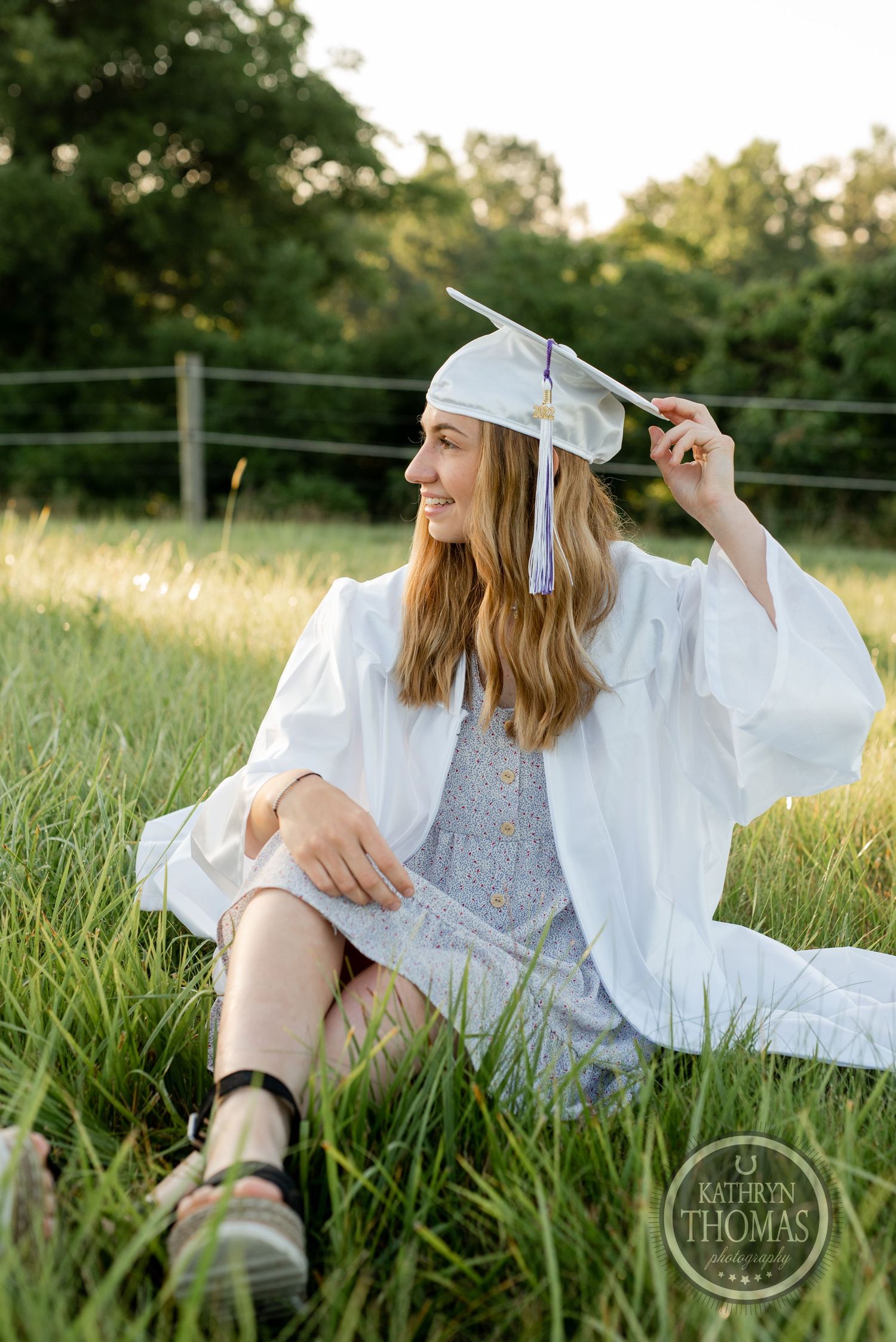 Graduate adjusts cap while sitting in tall grass during sunset graduation photo session.