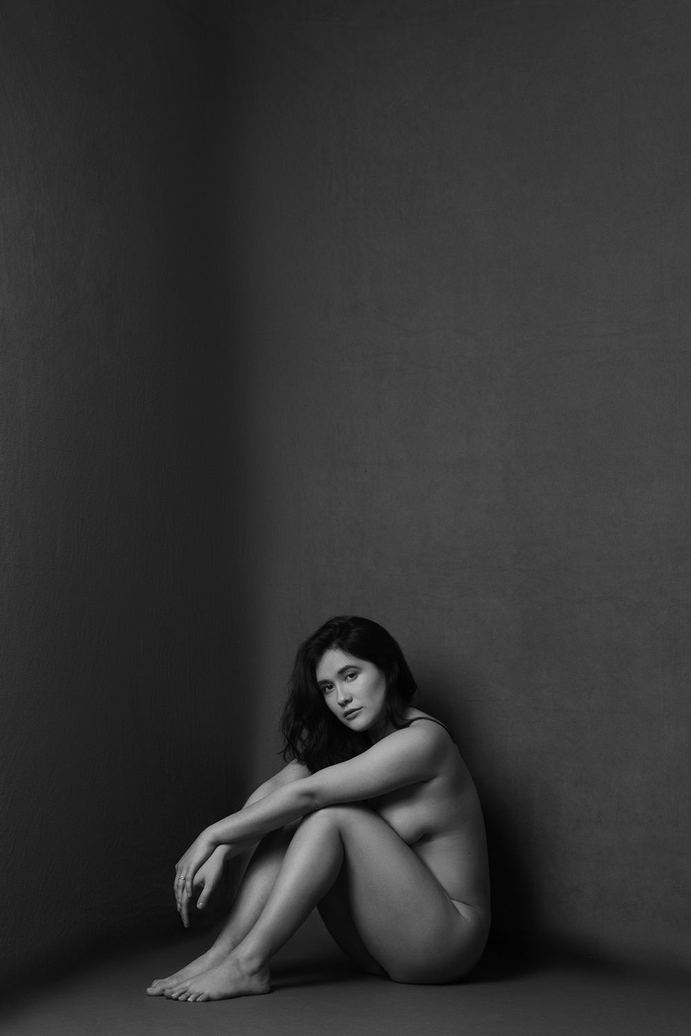 Minimalist nude study in corner setting with dramatic shadows and intimate pose.