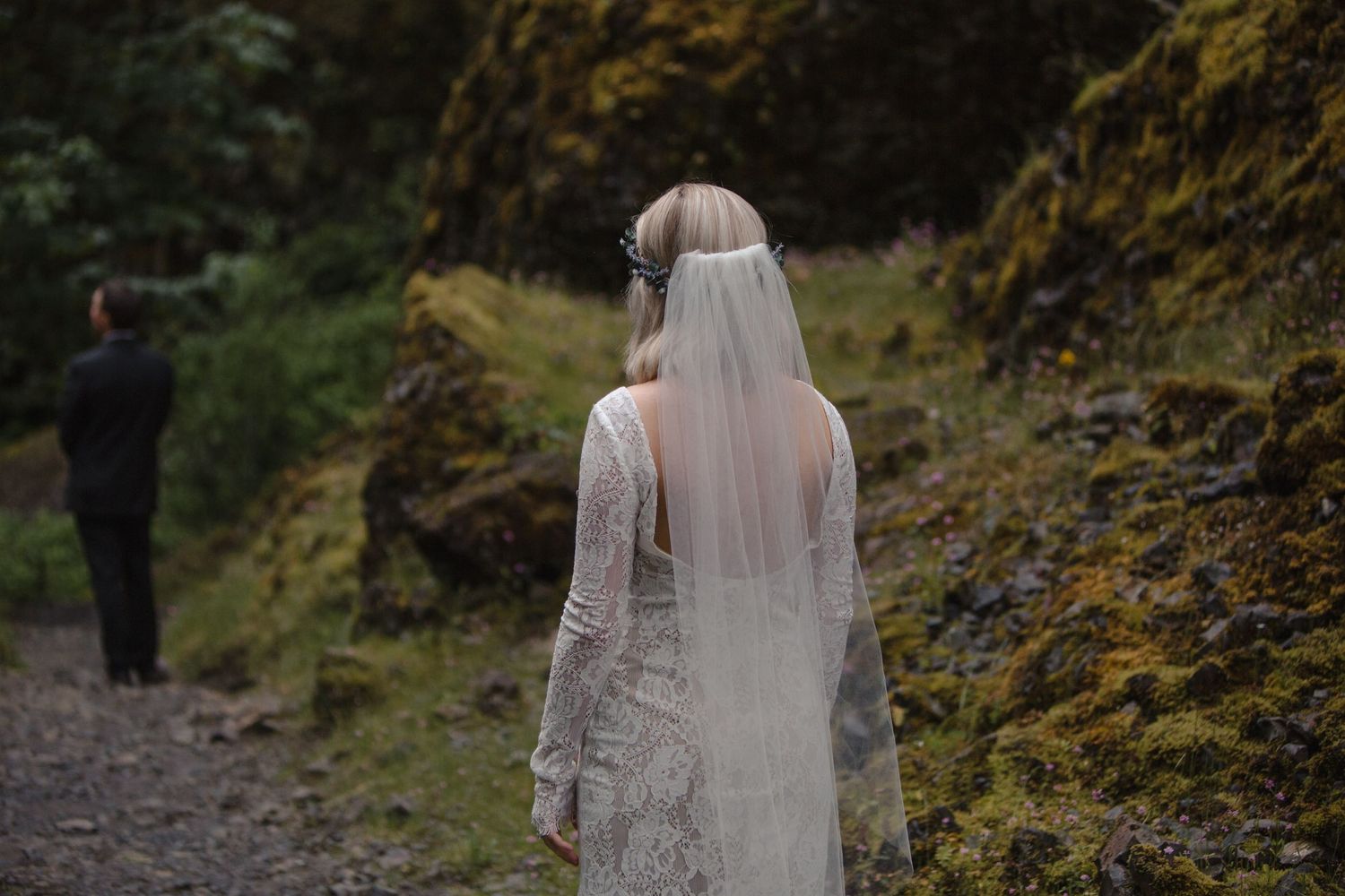 A bride in a flowing white dress and veil walking down a wooded forest path.