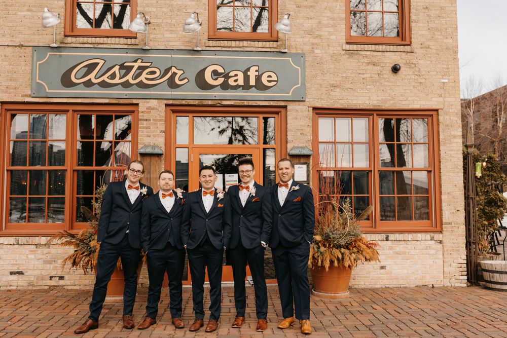Groom and groomsmen in navy suits with orange bow ties standing outside Aster Cafe in winter.