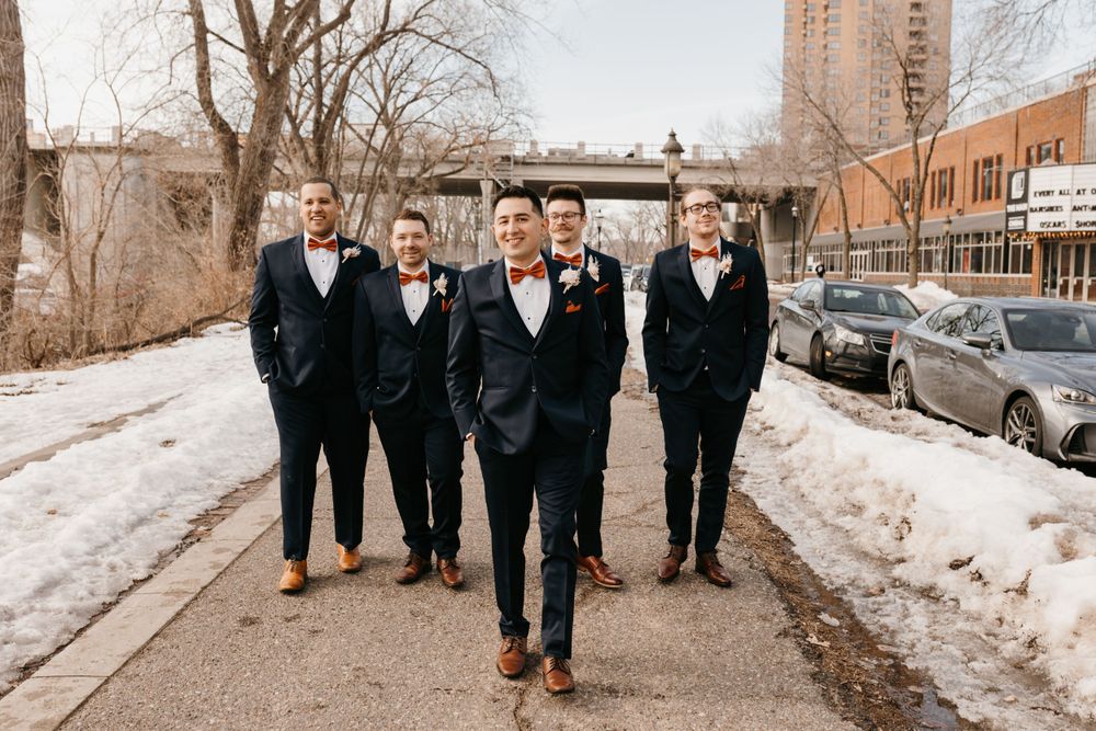 Groom and four groomsmen in navy suits with orange bow ties walking confidently on a snowy path.