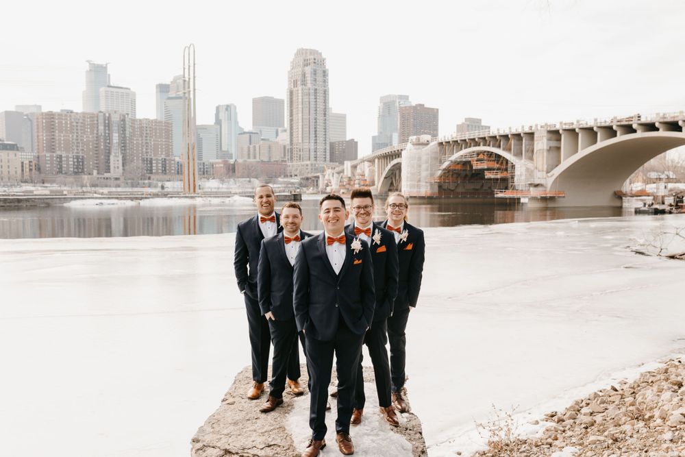 Groom and groomsmen in navy suits posing by a river with a city skyline and bridge backdrop.