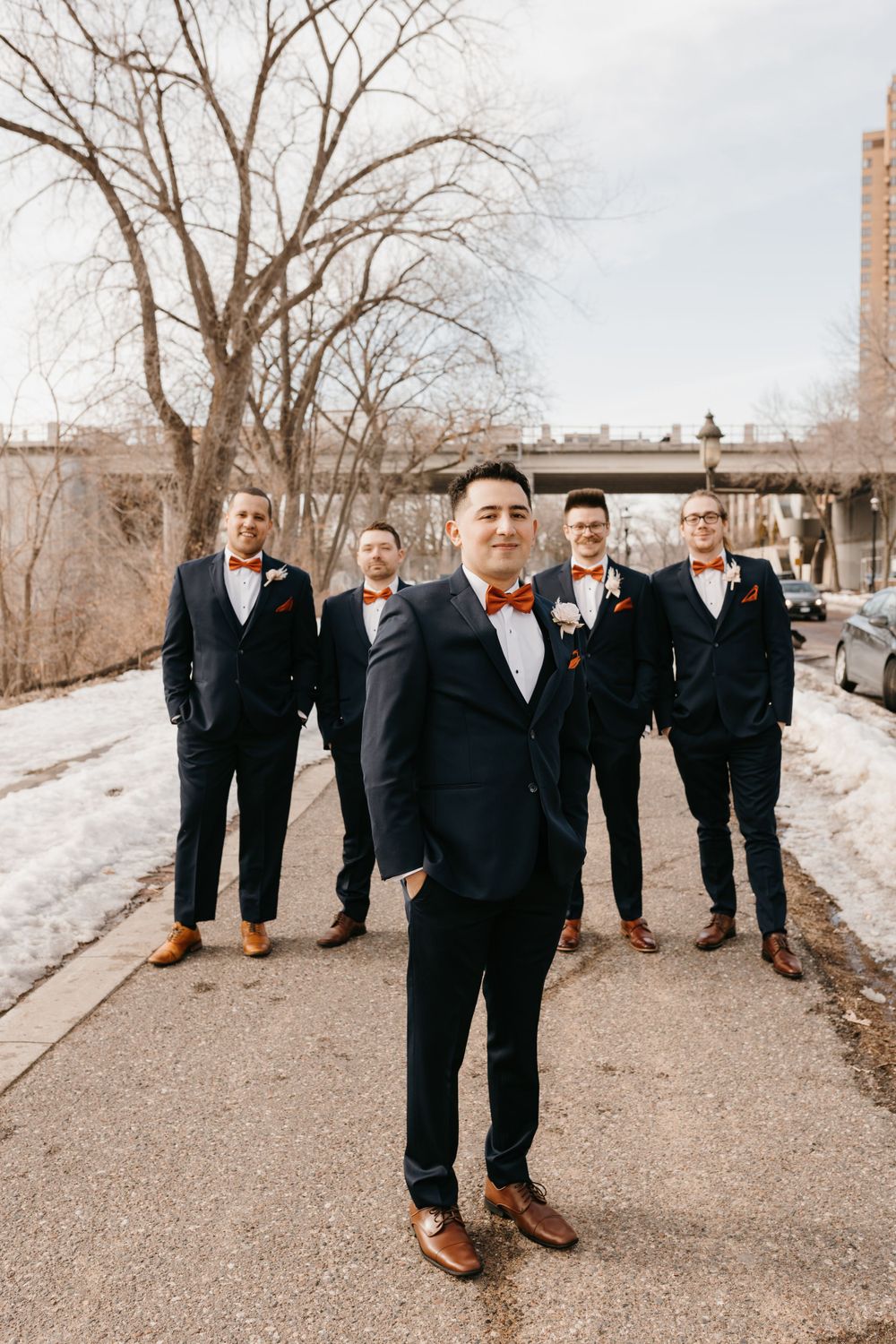 Groom in navy suit with orange bow tie stands in front of four groomsmen on a winter pathway.