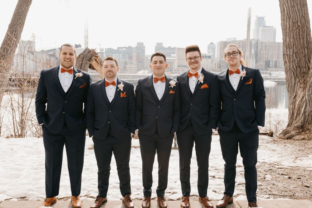 Five groomsmen in navy suits with orange bow ties posing outdoors with winter city skyline backdrop.