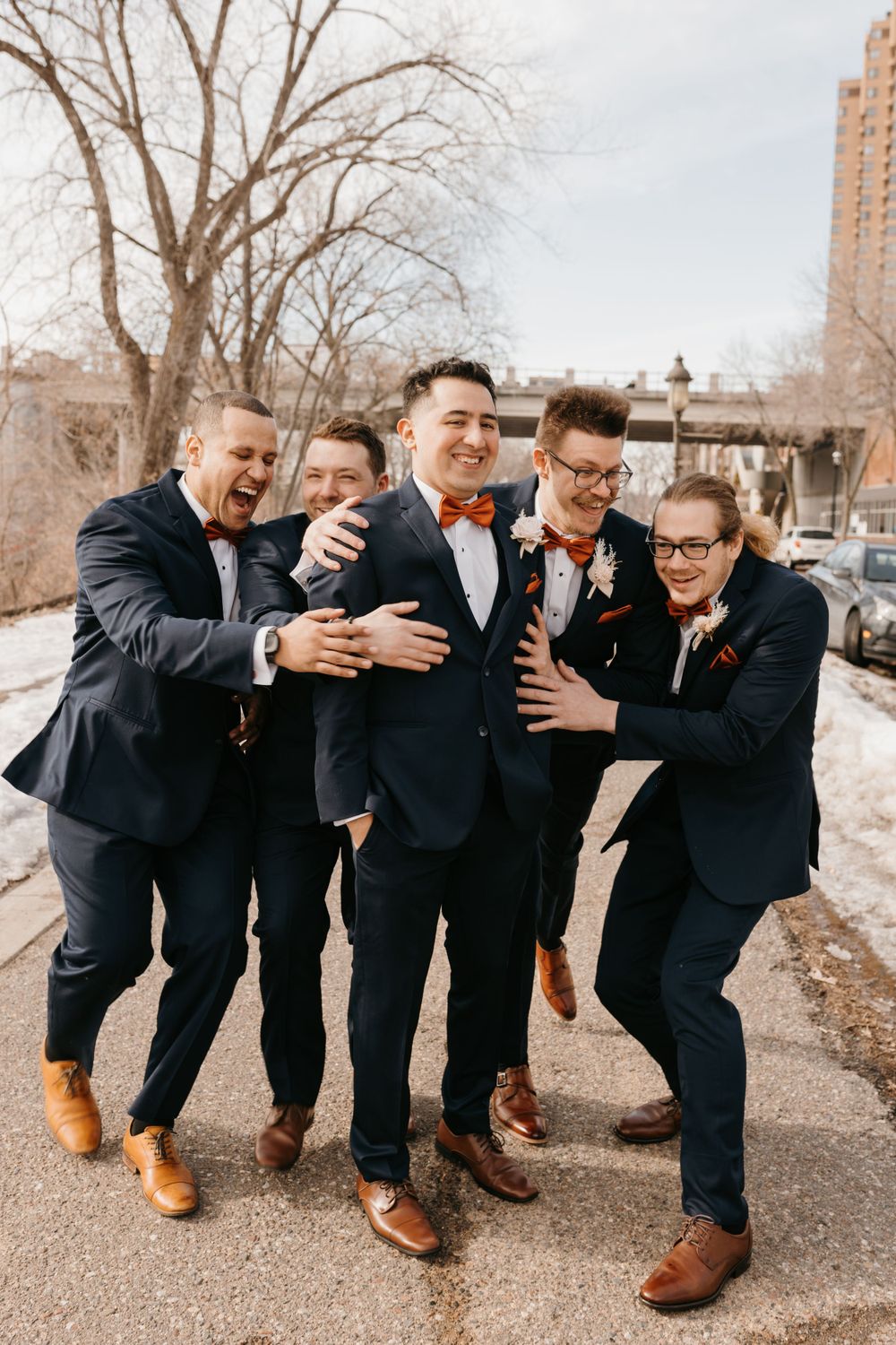 Groom laughing as four groomsmen playfully crowd around him in navy suits with orange bow ties.