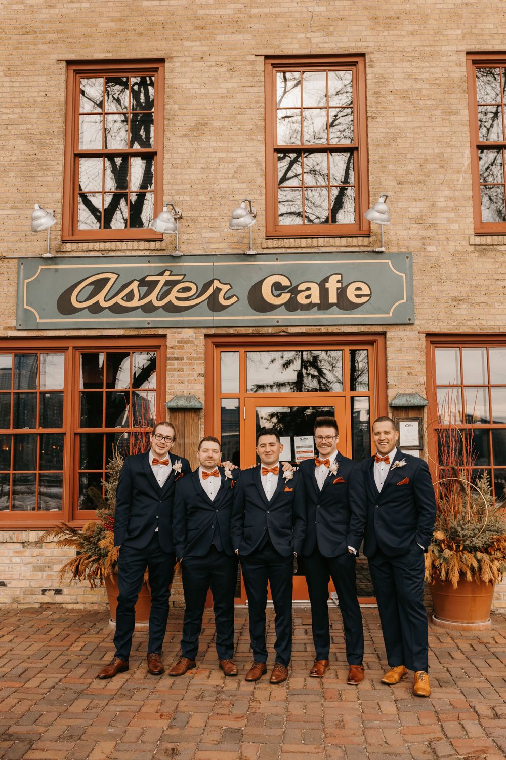 Five groomsmen in navy suits with orange bow ties posing in front of Aster Cafe brick building.