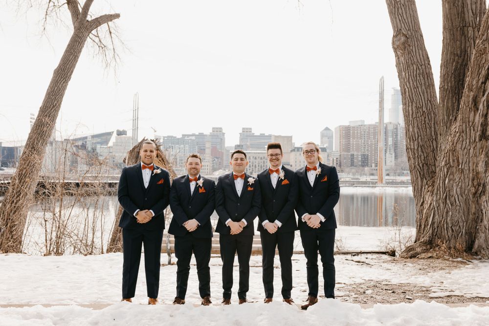Groom and four groomsmen in navy suits with bow ties standing in snow with city skyline behind.