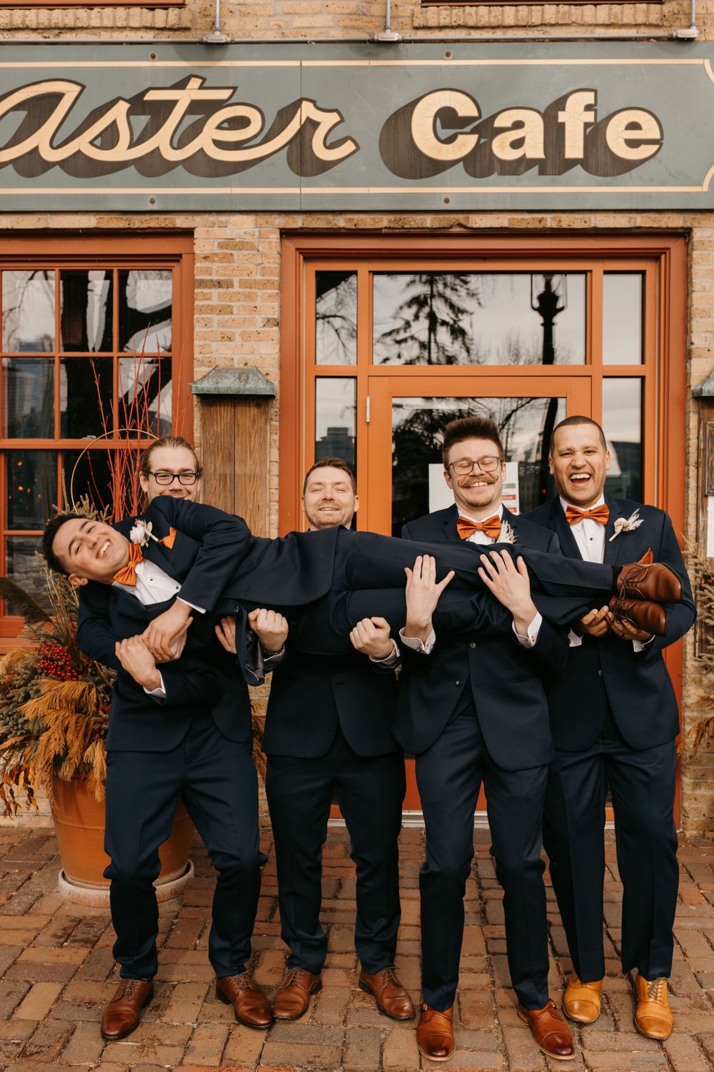 Groomsmen humorously lifting the groom outside Aster Cafe while wearing navy suits and orange bow ties.