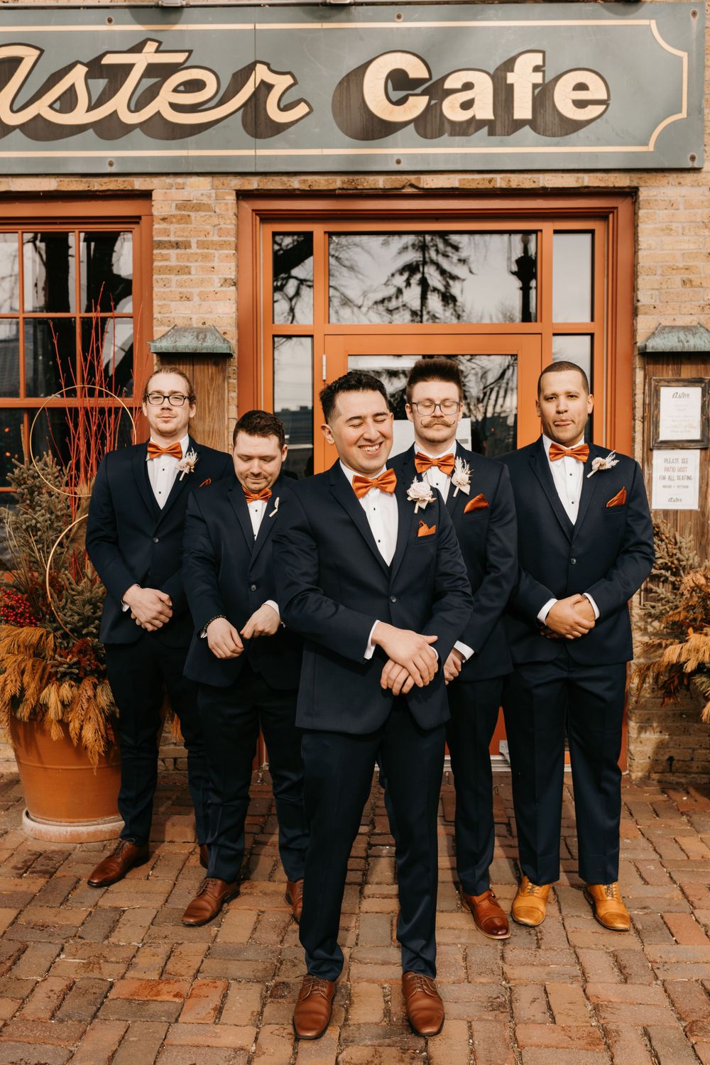 Groom smiling confidently in front of groomsmen outside Aster Cafe in navy suits and orange bow ties.