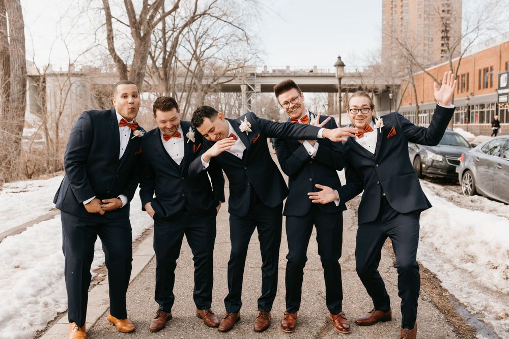 Five groomsmen in navy suits with orange bow ties laughing and posing playfully outdoors in winter.