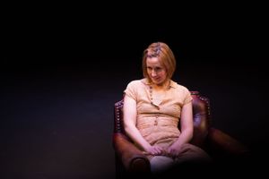 A dimly lit scene showing someone seated in a dark theater or auditorium setting.