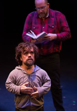 A performer in a gray sweatshirt gesturing during what appears to be a theatrical scene.