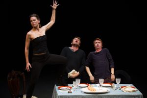 Performers enact a dramatic scene around a dining table on a darkened stage.