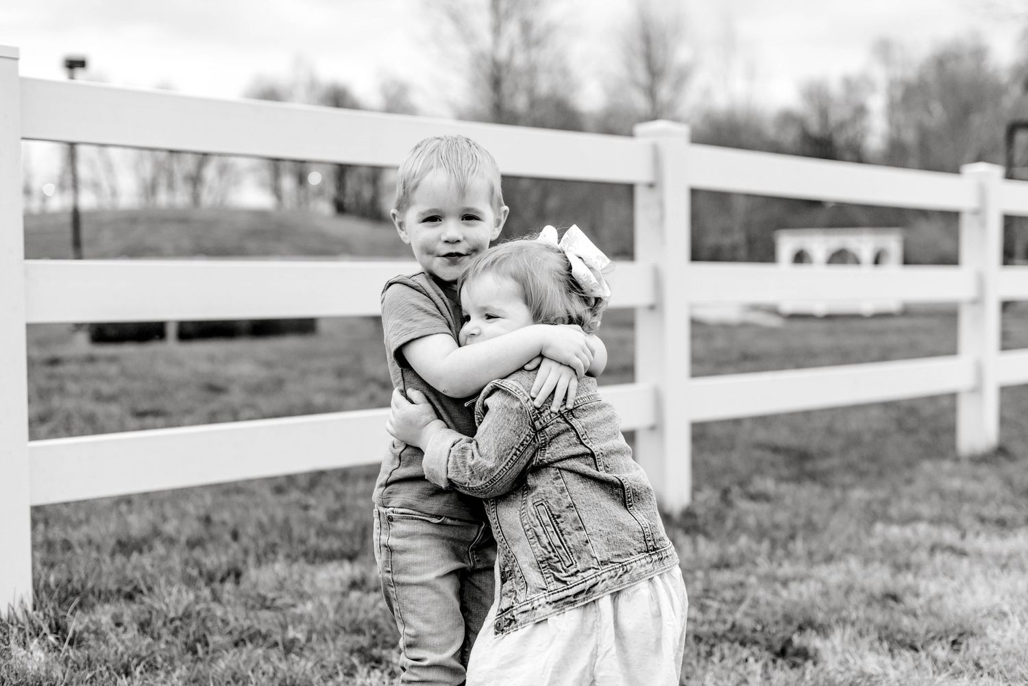Two young children embrace in a sweet hug by a white fence in black and white.