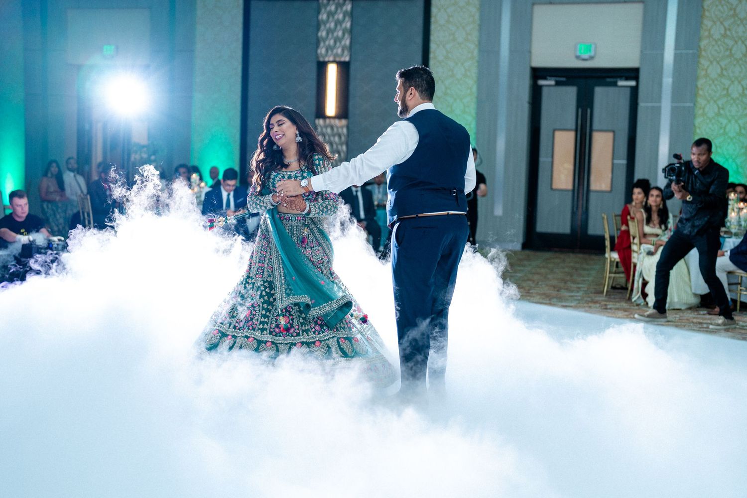 Couple twirls amidst fog's mystique, enveloped in ethereal green and blue hues, a captivating celebration of love.