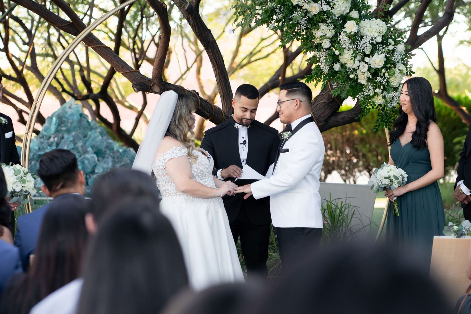 Outdoor wedding ceremony under trees with guests watching the exchange of vows.