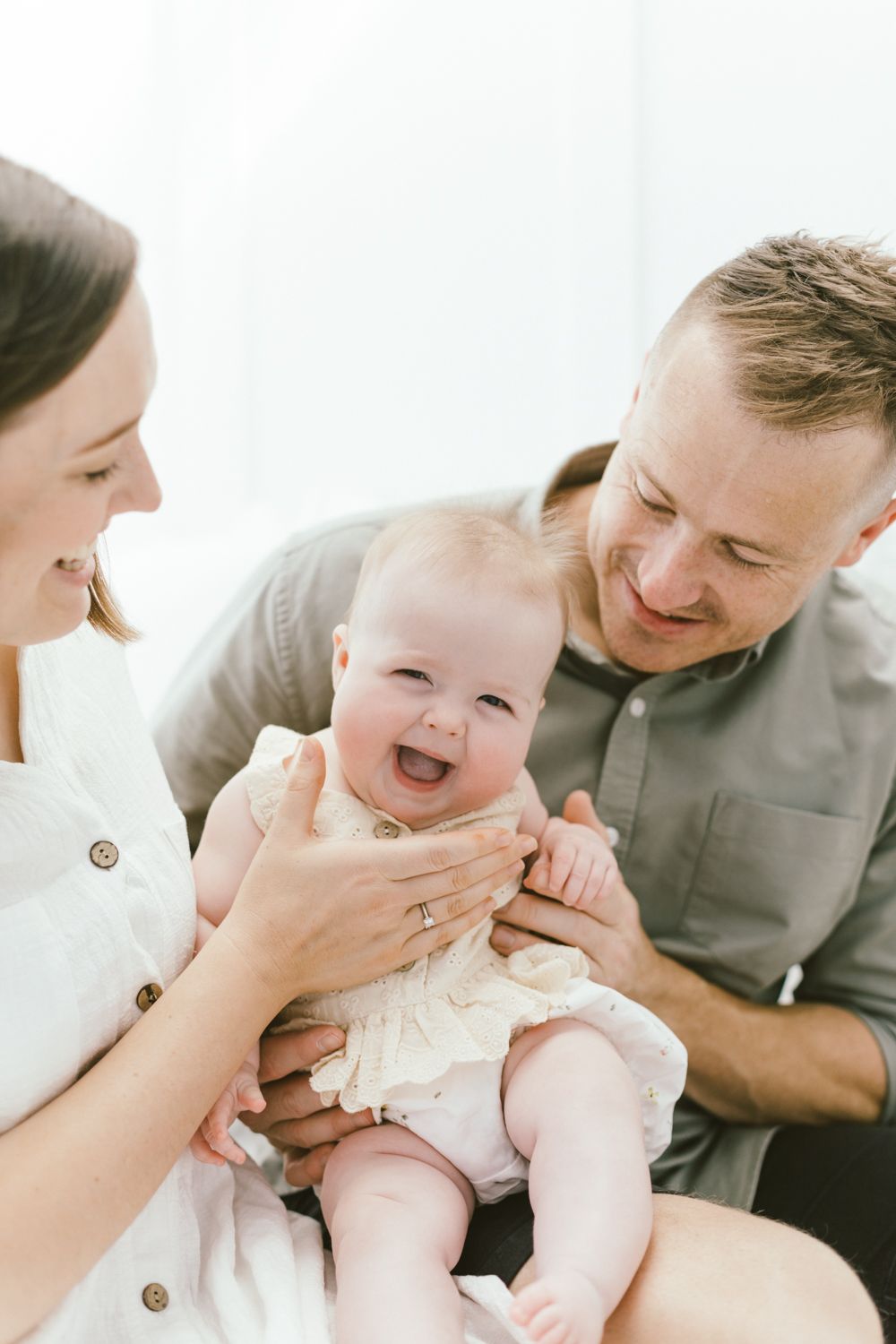 Candid family portrait showing playful interaction with laughing baby in cream outfit.