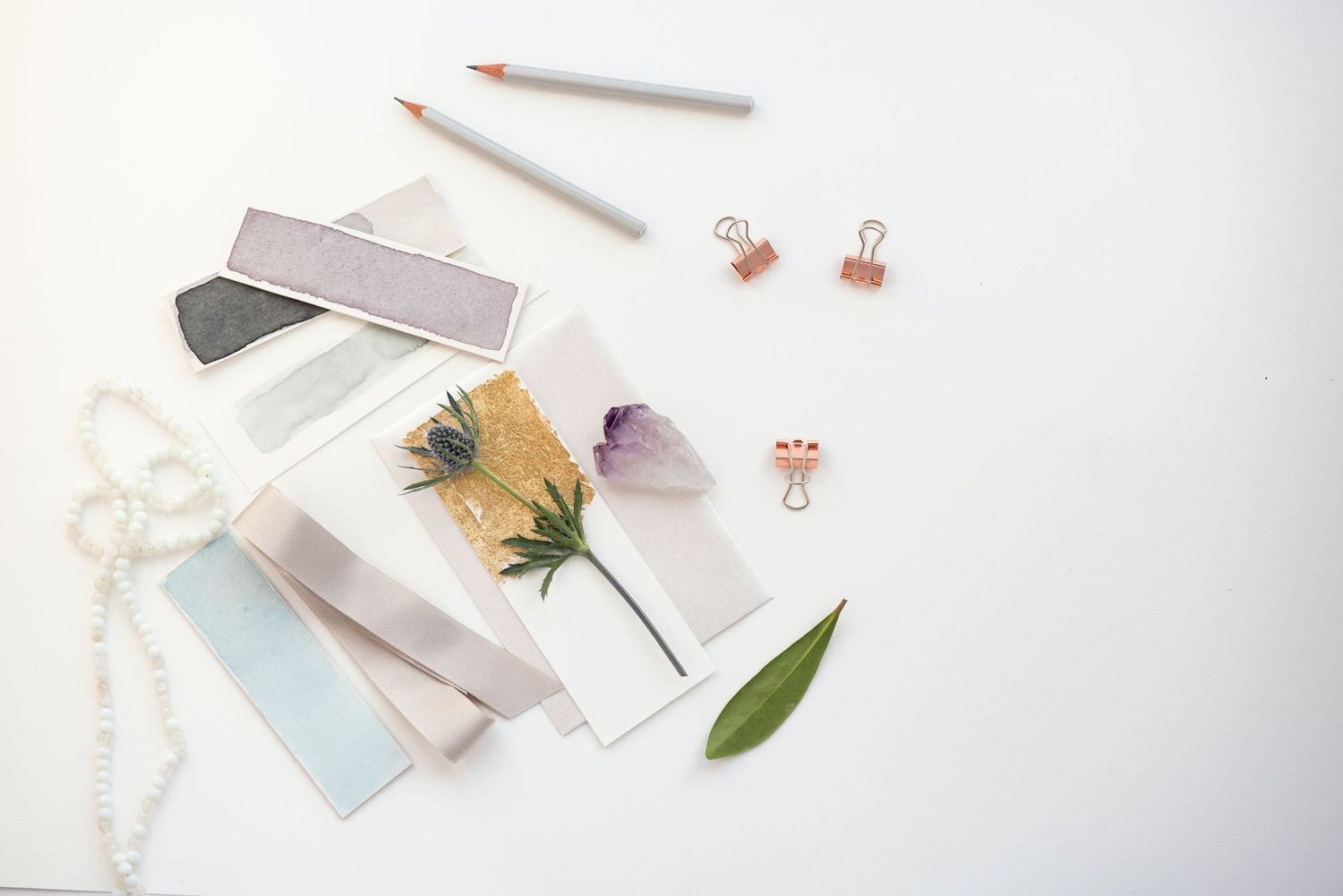 Flatlay of stationery items including pencils, dried flowers, and rose gold accessories on white background.