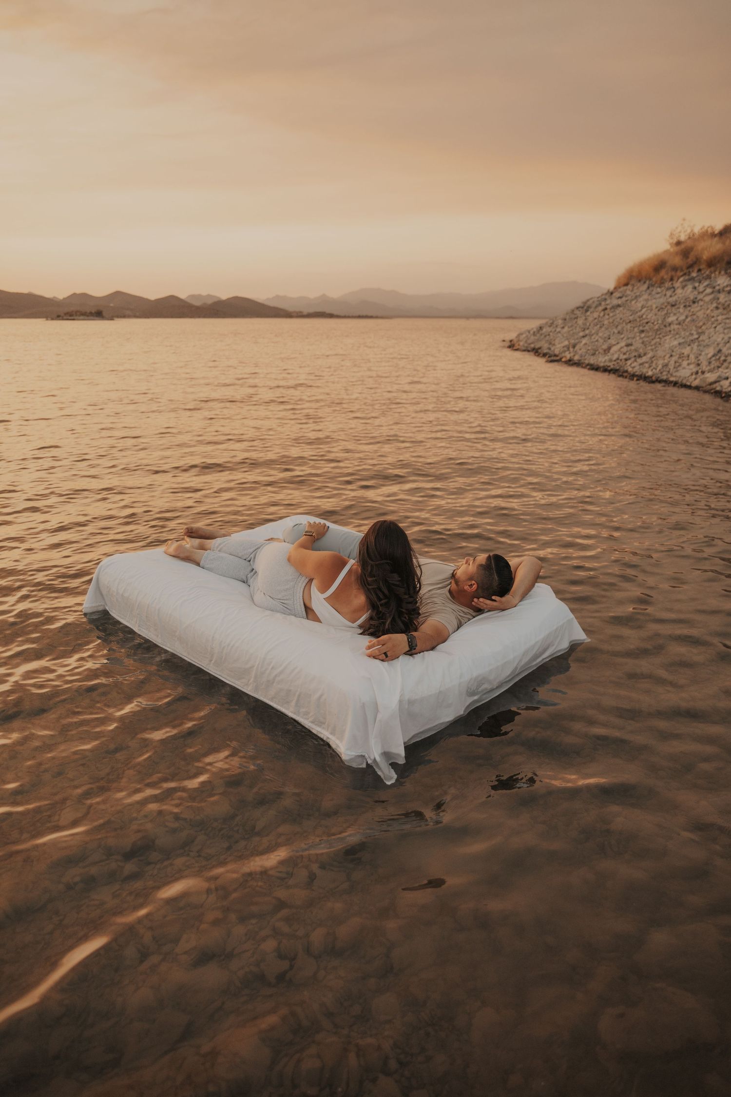 casually dresses couple lounging on an air mattress that floats on a lake overlooking the sunset