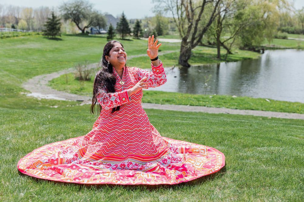 Woman in flowing pink dress sitting on grass near pond taking selfies or photos.