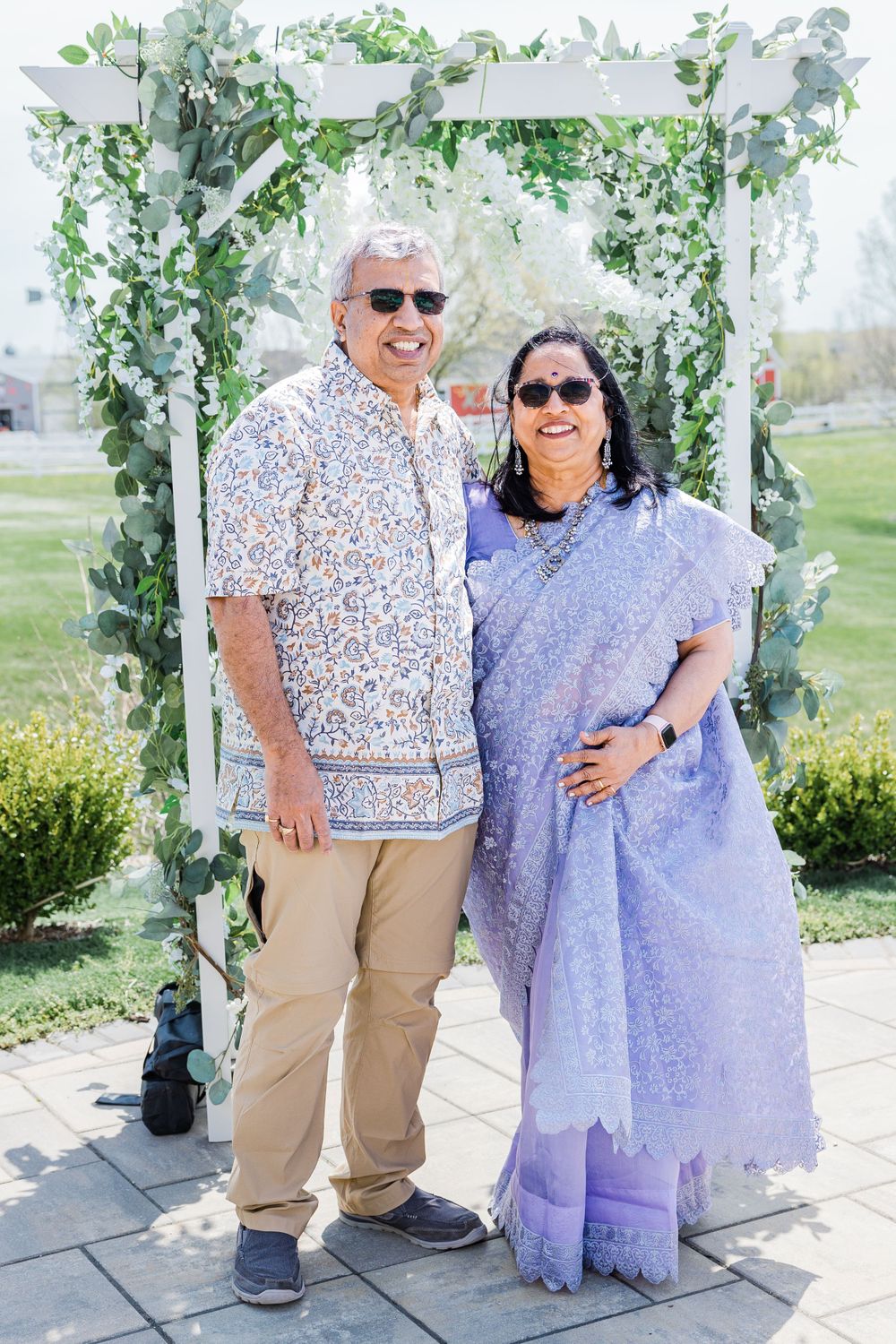 A couple poses under a garden arch decorated with greenery, wearing casual and traditional Indian attire.