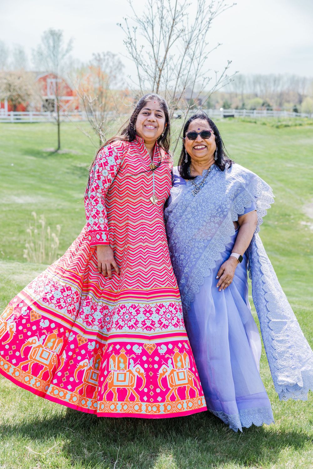 Two women pose together outdoors, wearing blue sari and vibrant pink traditional dress.