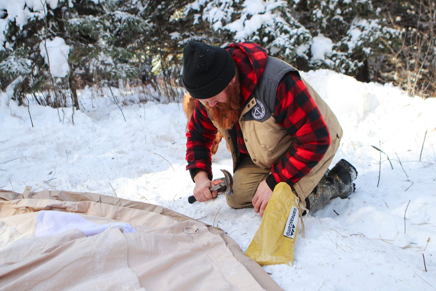 Person in buffalo plaid jacket setting up camping equipment in snow covered woods.