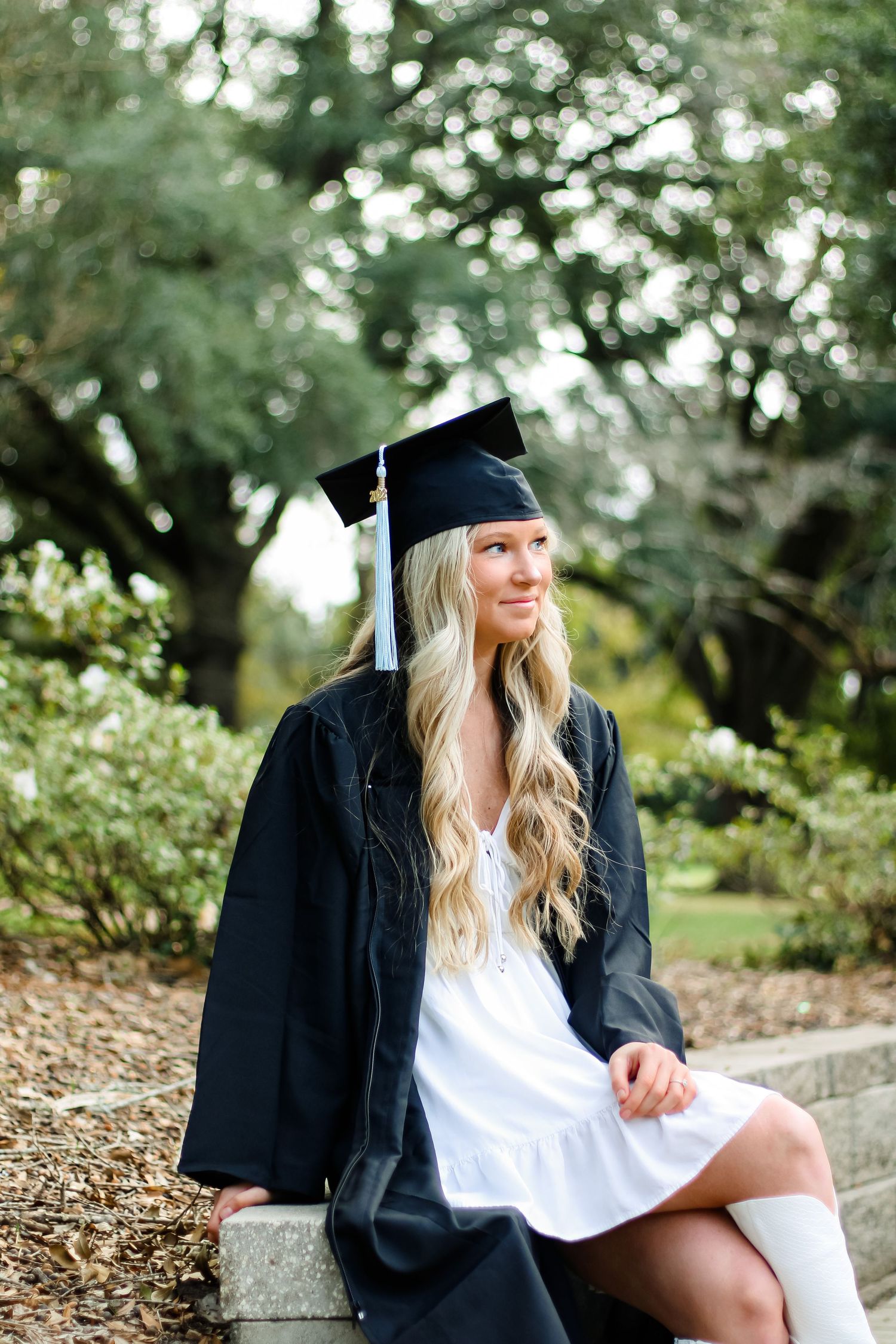 Graduate in traditional cap and gown sits thoughtfully in a garden setting.