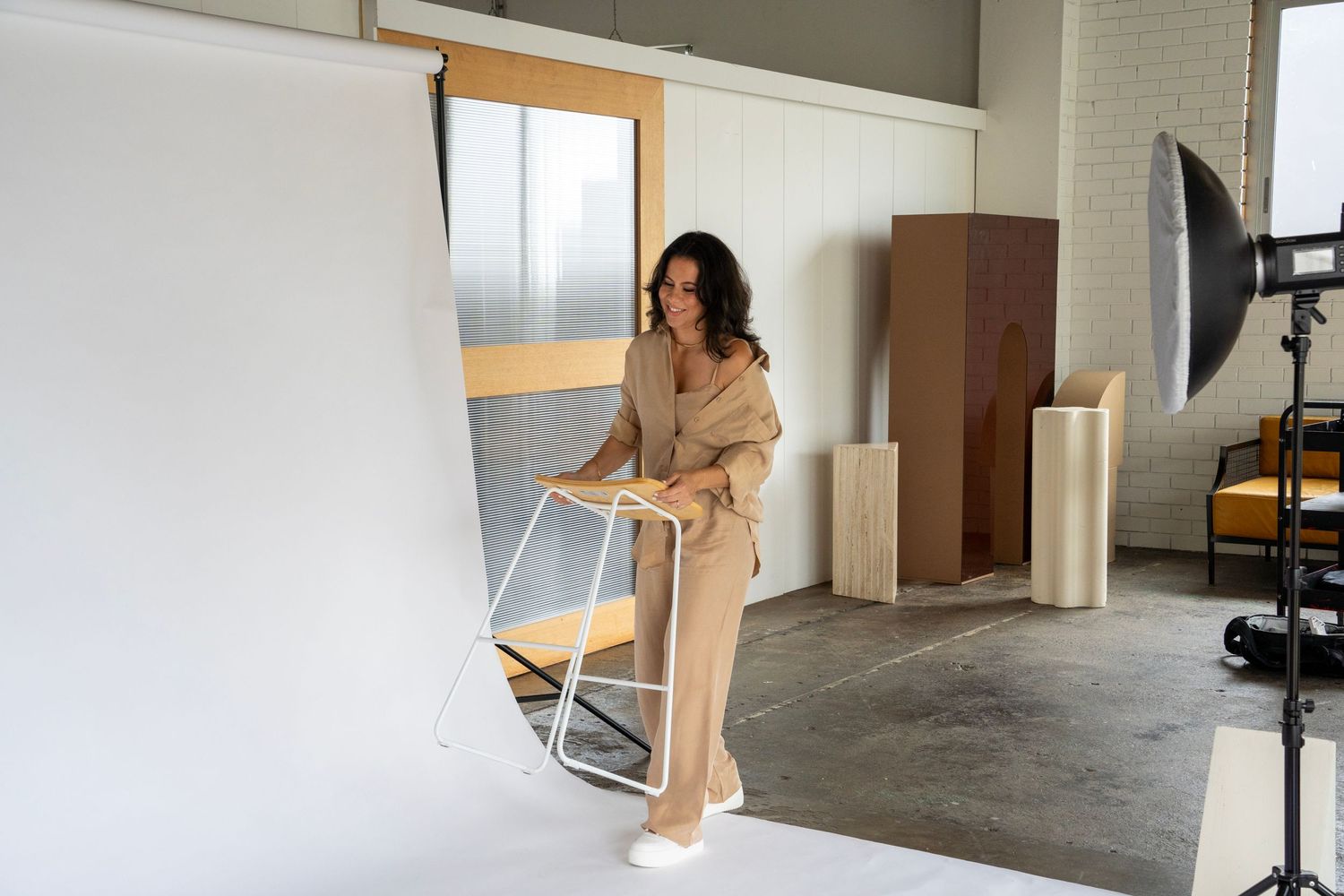 A photographer in a beige outfit arranges studio equipment near a white backdrop.