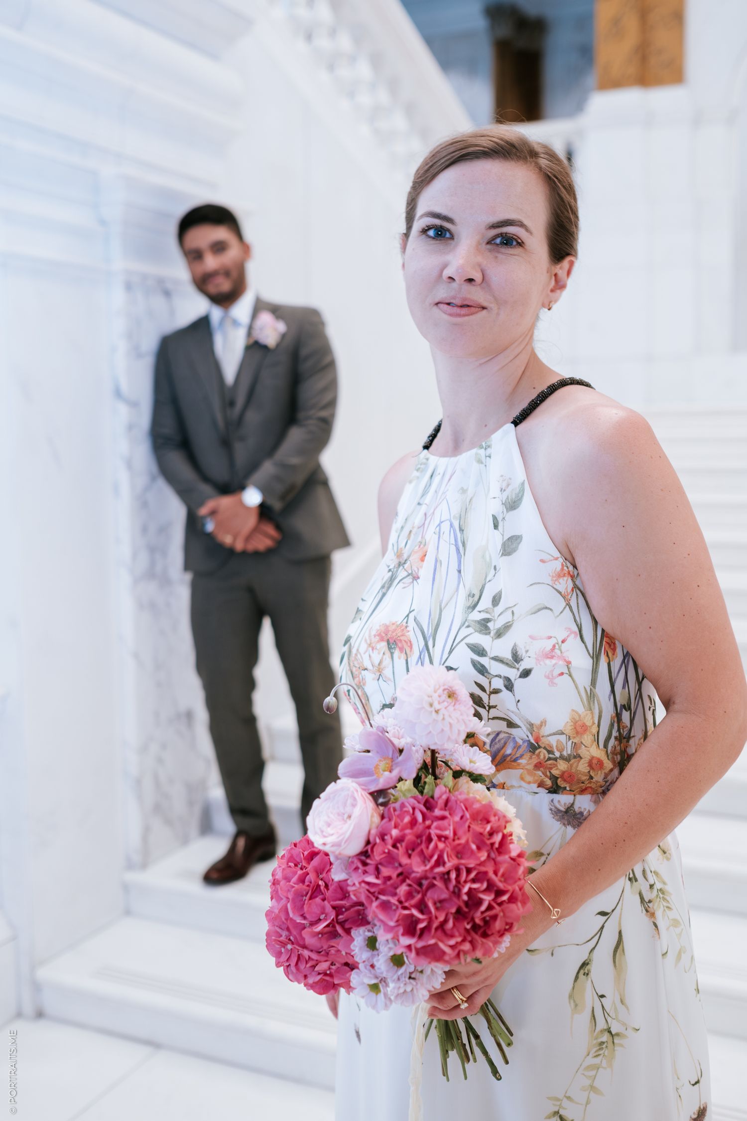 A person in a floral dress holds a vibrant pink bouquet against a white architectural backdrop.