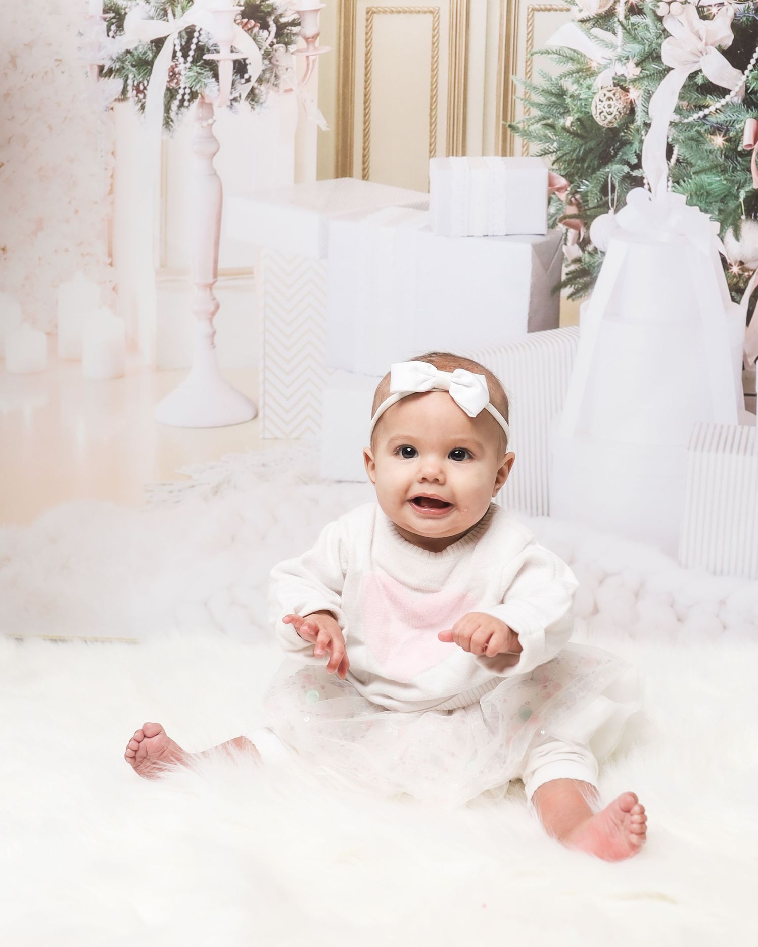 Baby wearing a white dress withbow headband sits on soft white blanket surrounded by holiday decor.