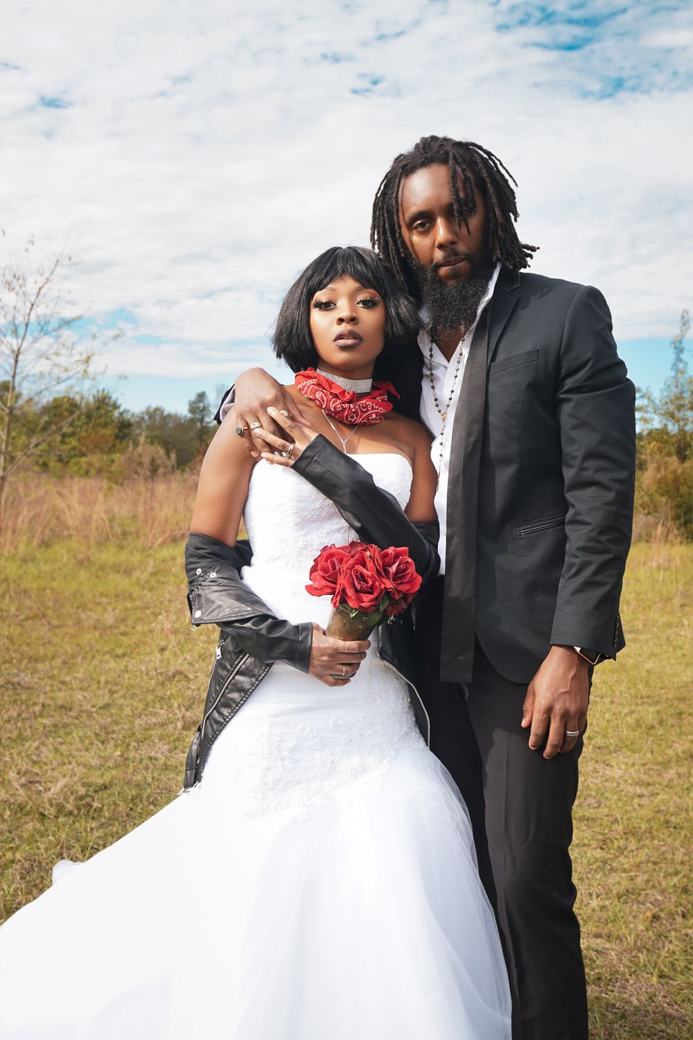 Wedding photo with bride in white gown with red accents and groom in black suit outdoors.