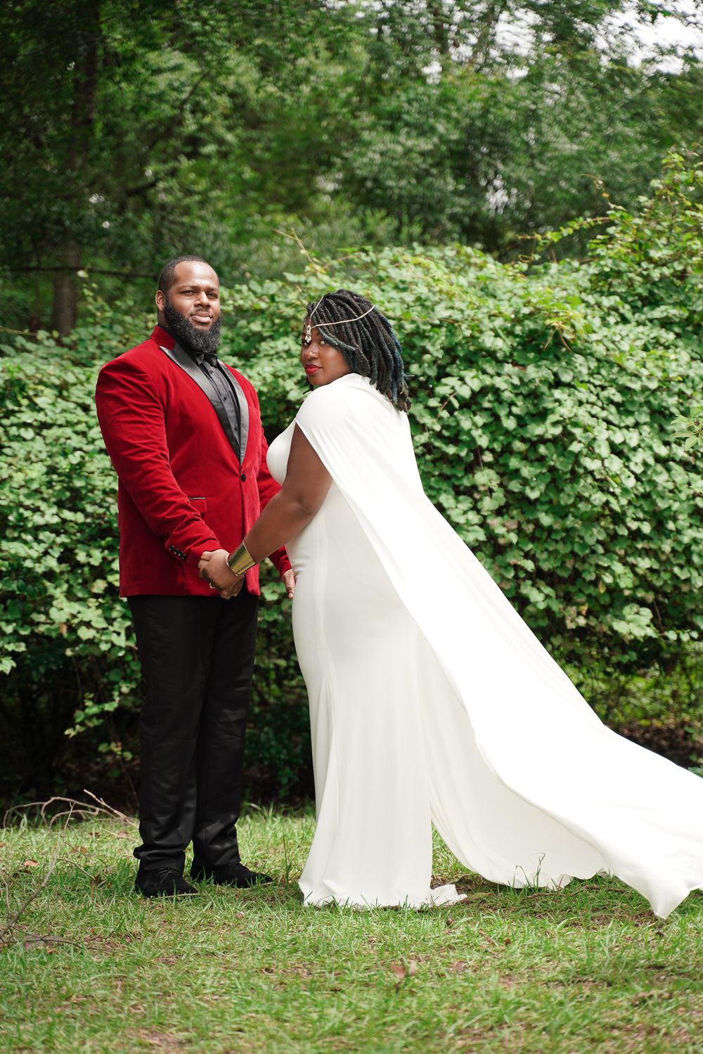 A wedding couple in a red blazer and flowing white dress pose together in a garden setting.