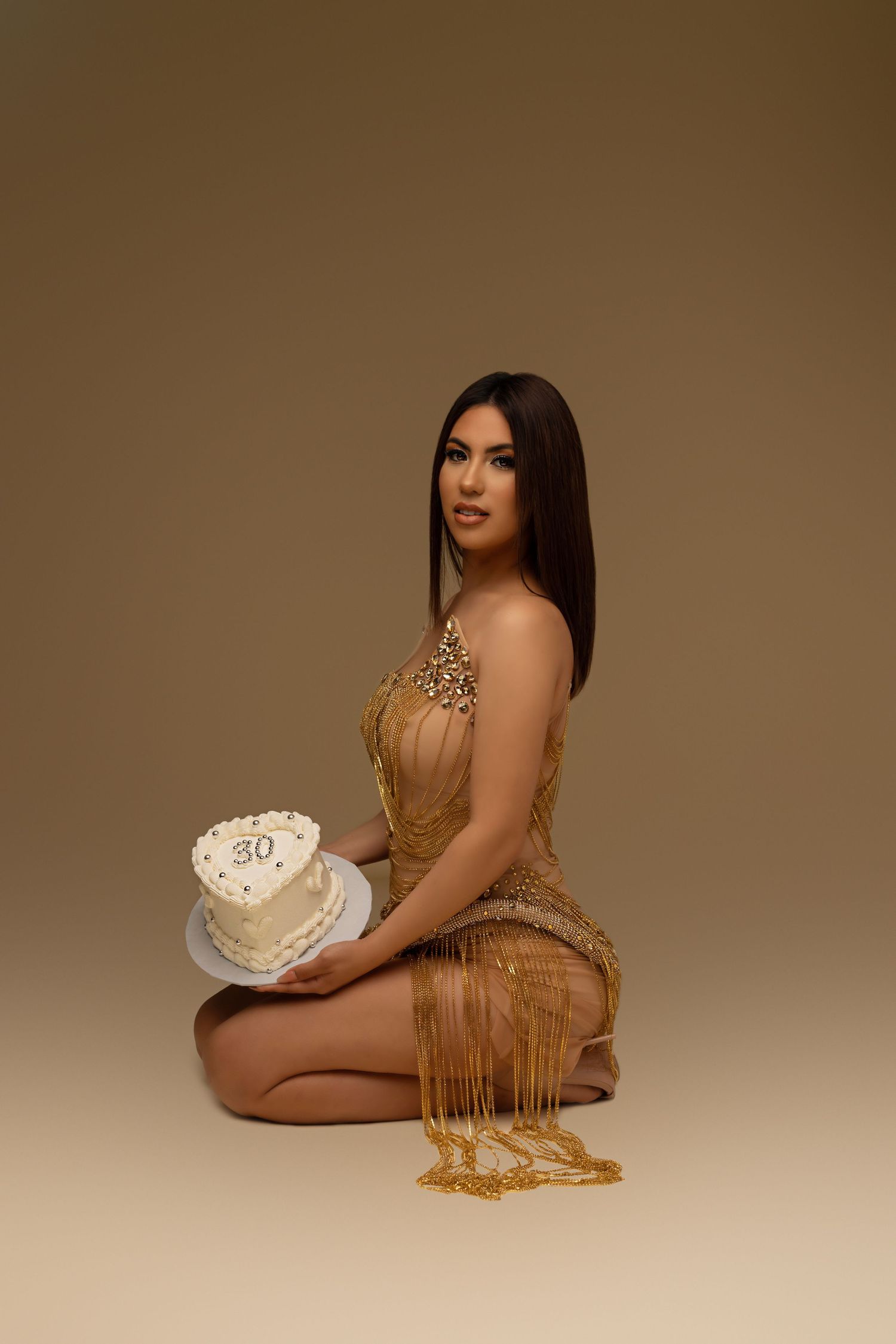 Glamorous photoshoot featuring a gold fringed dress and white birthday cake against a brown backdrop.