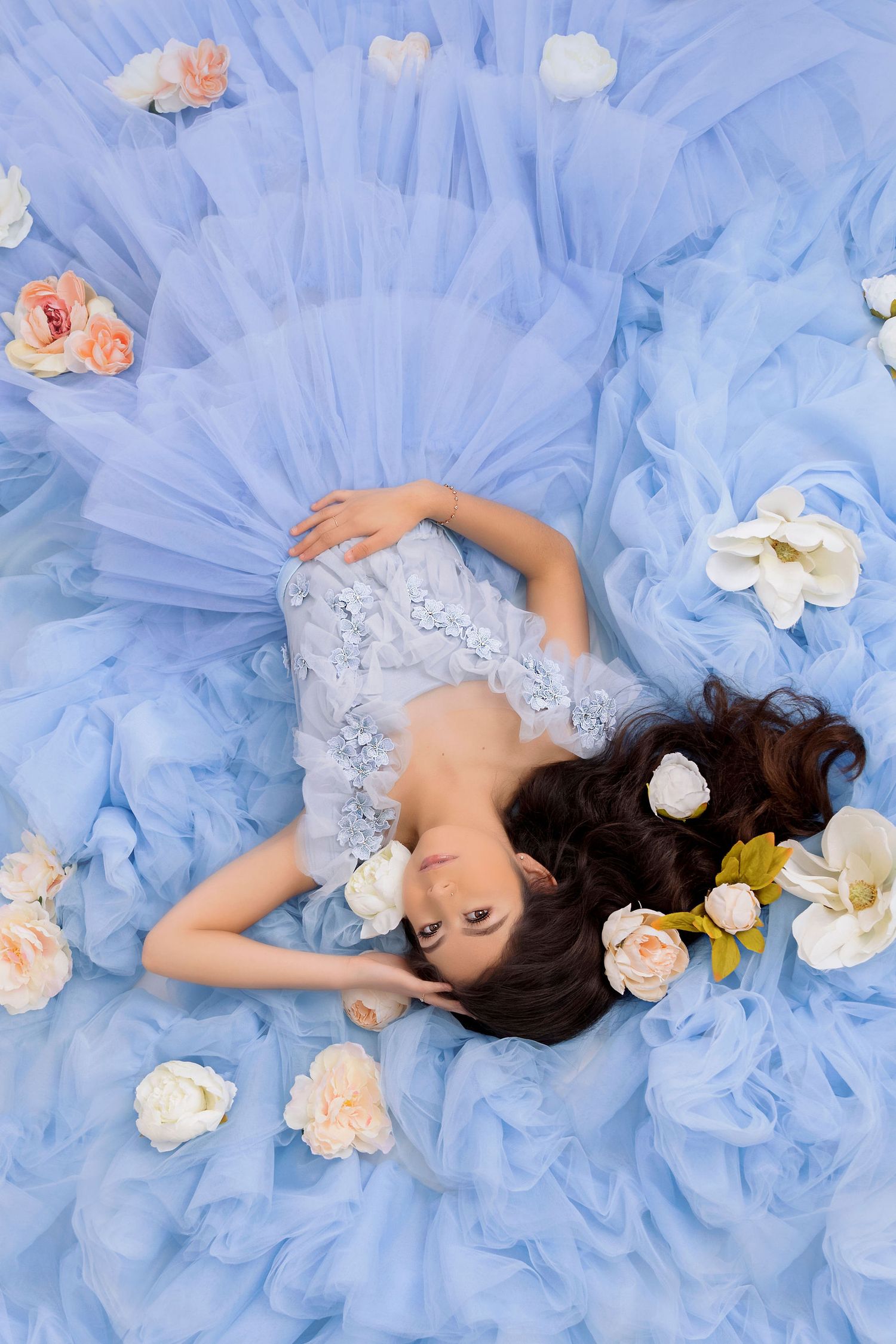 Ethereal portrait of a figure lying on blue tulle fabric surrounded by scattered pink and white roses.