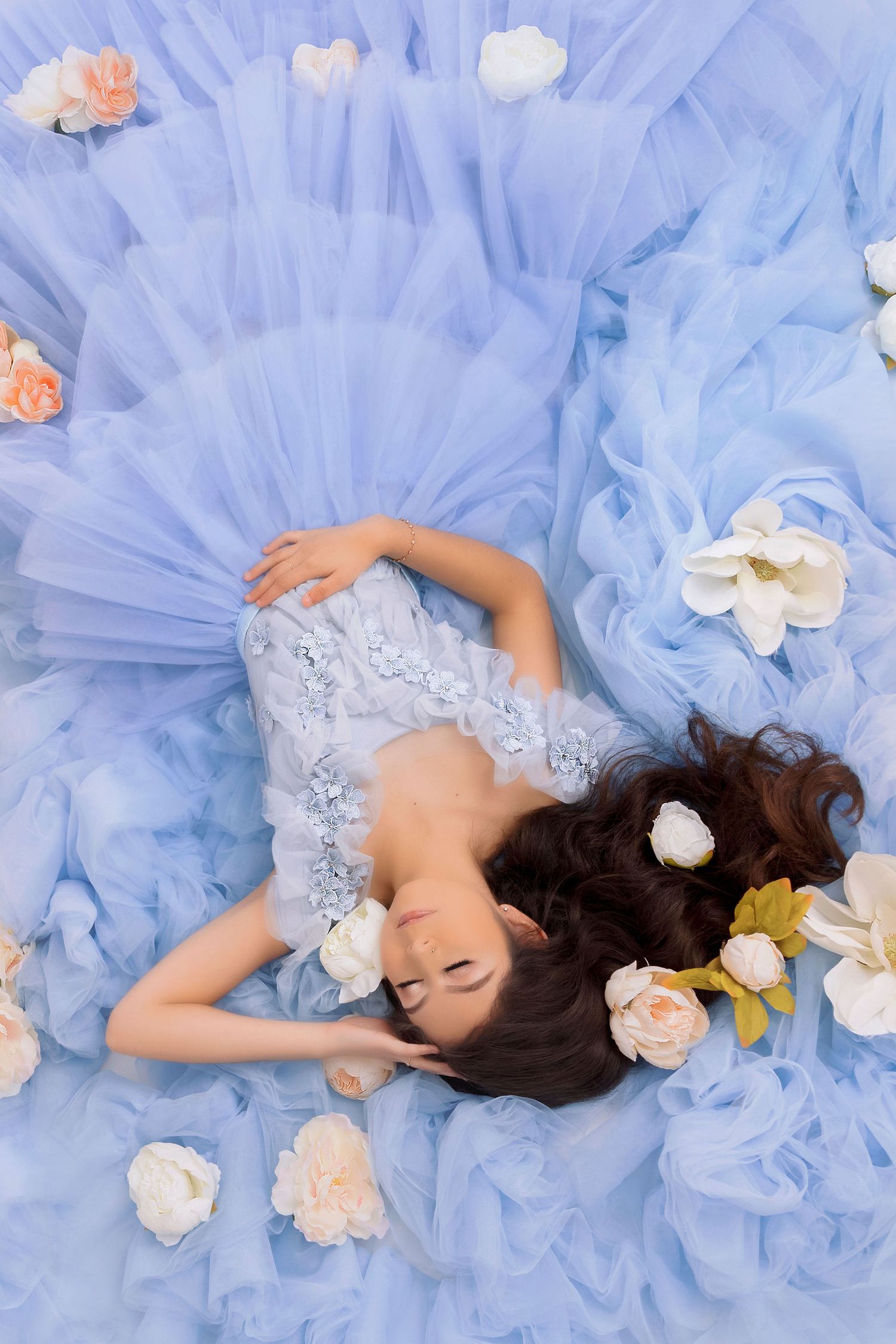 Overhead shot of woman in blue tulle gown lying among scattered flowers.