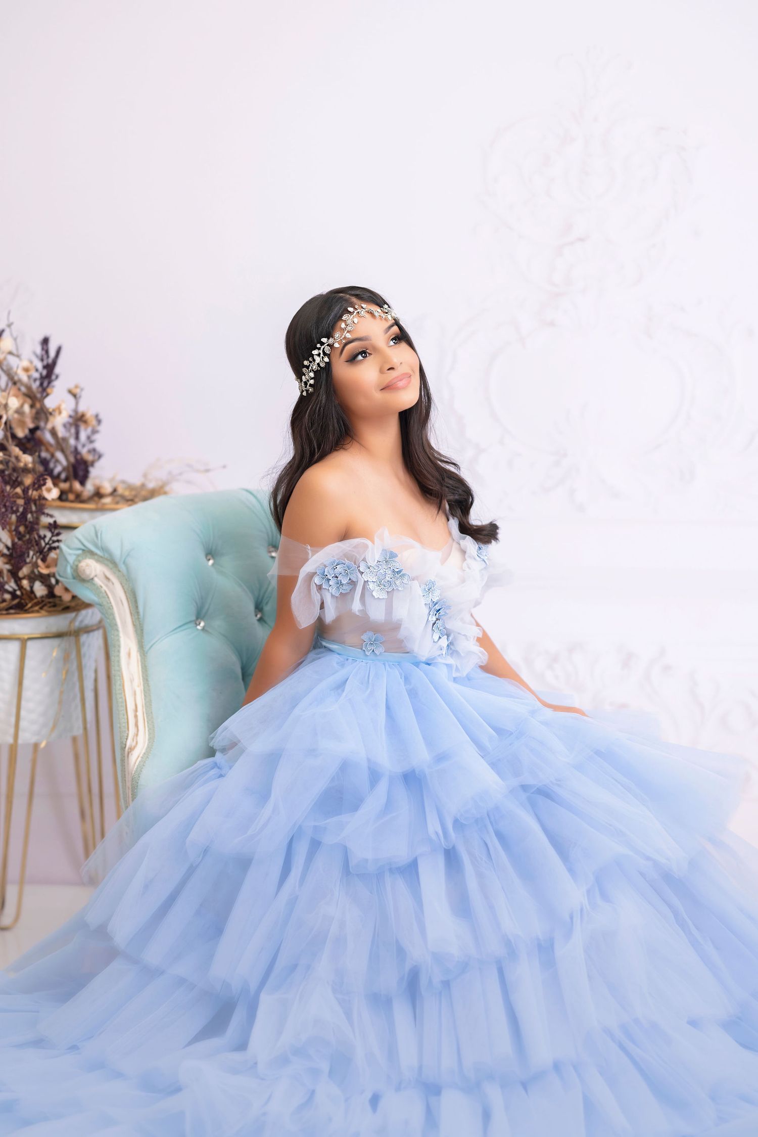 Woman in layered blue tulle ballgown poses on tufted chair with crystal headpiece.