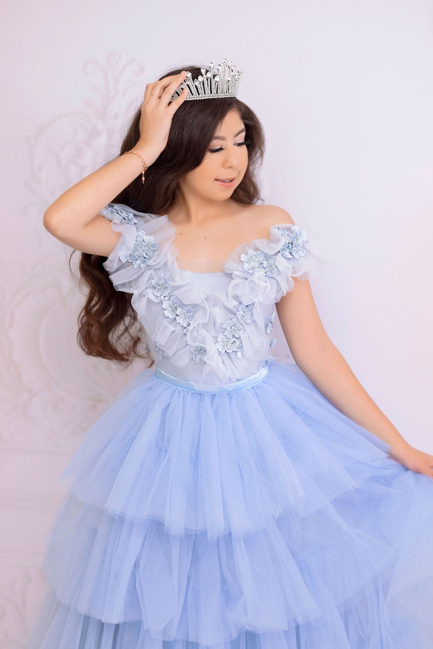 Woman wearing light blue ruffled gown adjusts crystal tiara in romantic portrait setting.